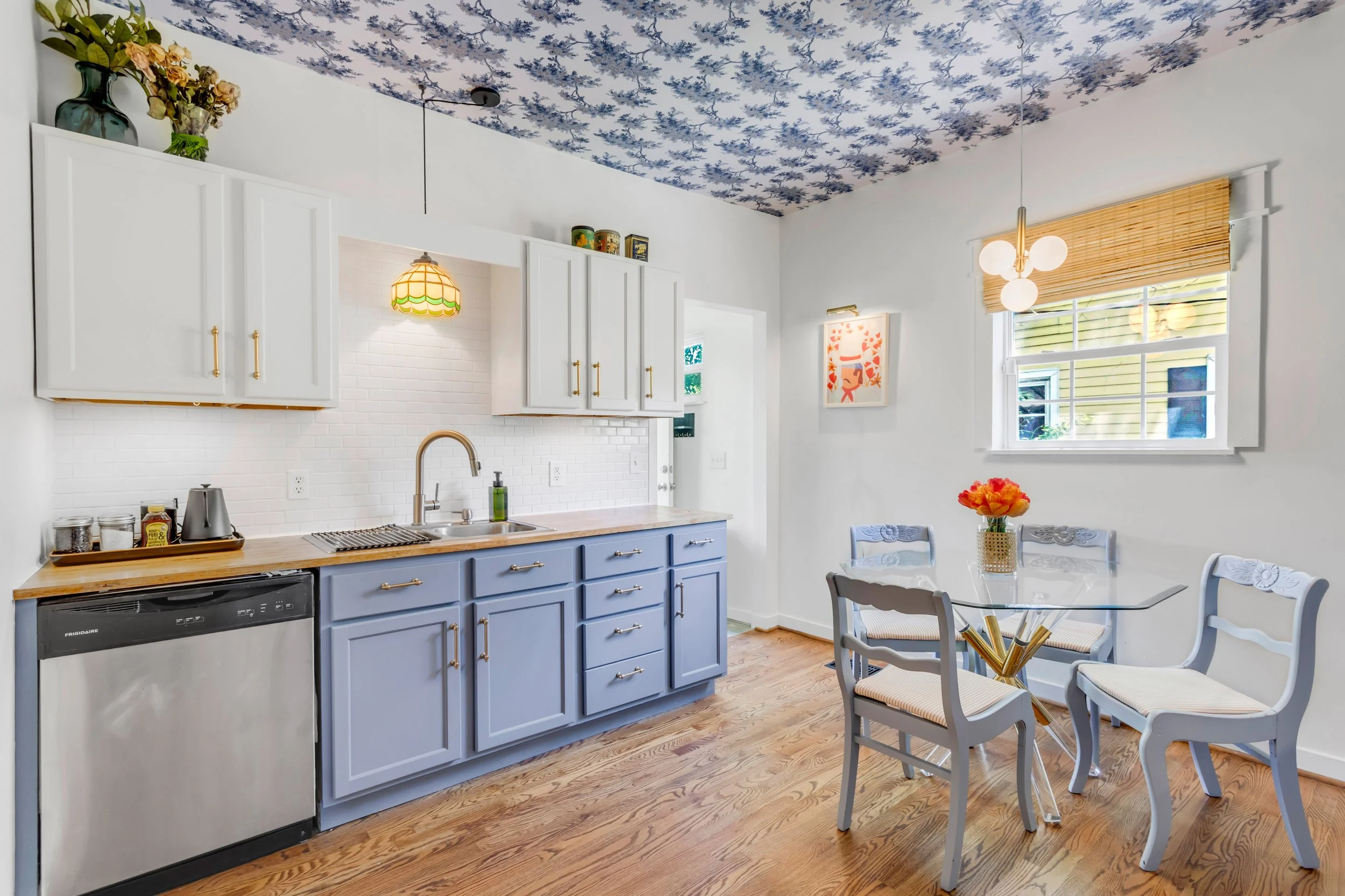 A brightly lit kitchen dining area with white walls, a white brick backsplash, and a ceiling with blue floral wallpaper pattern. Cabinetry is painted light blue with gold handles. There is a window with a bamboo shade. A white round dining table with