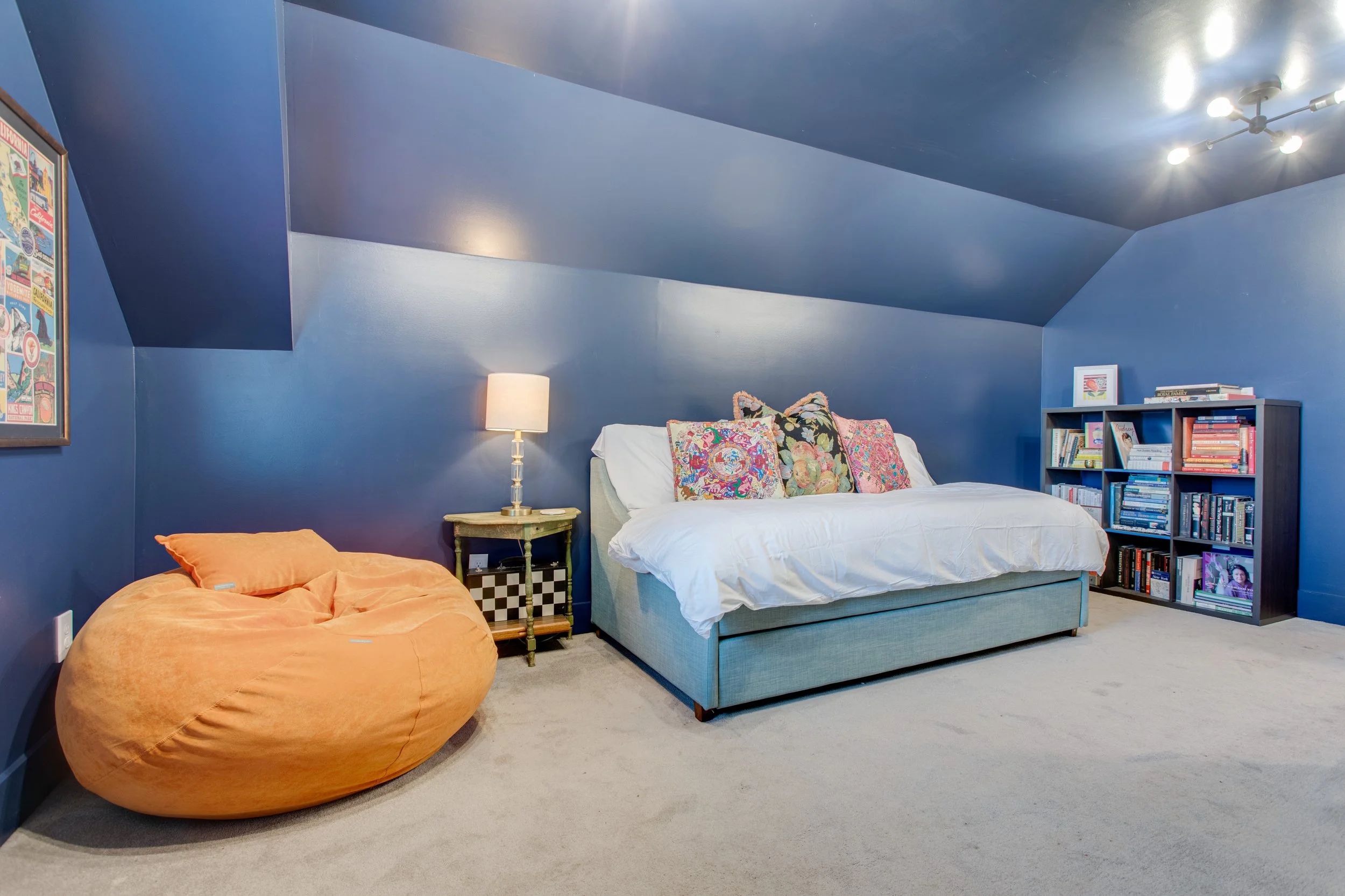 A bedroom with blue walls and ceiling, featuring a white bed with colorful embroidered pillows, a small wooden side table with a lamp, an orange bean bag chair, a bookshelf filled with books, a framed picture, and a colorful poster on the wall. A cei