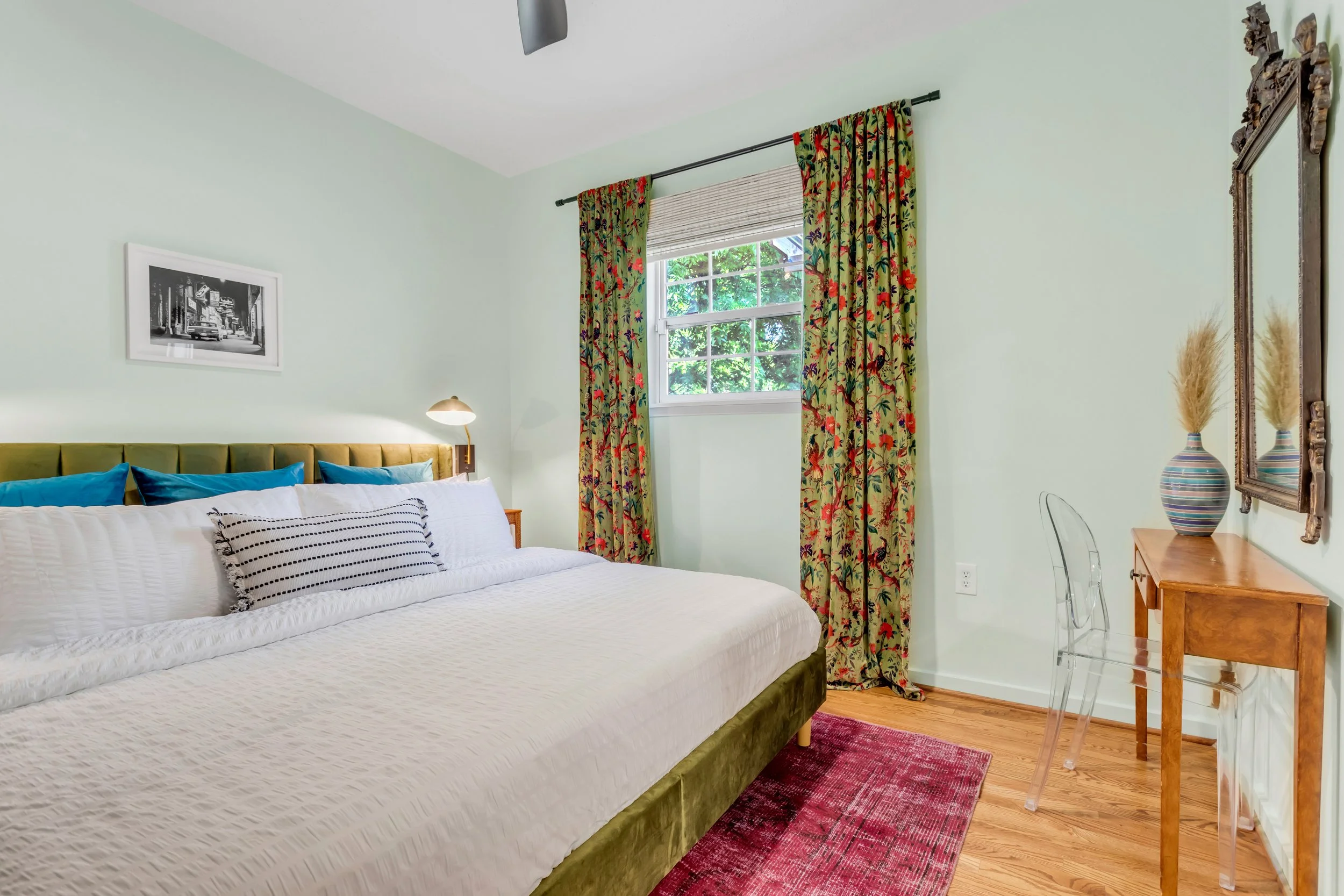 A bedroom featuring a large bed with white bedding and multiple blue pillows. There is a bedside lamp and a framed black-and-white picture on the wall. The room has light green walls, a window with floral curtains, and a wooden desk with a transparen