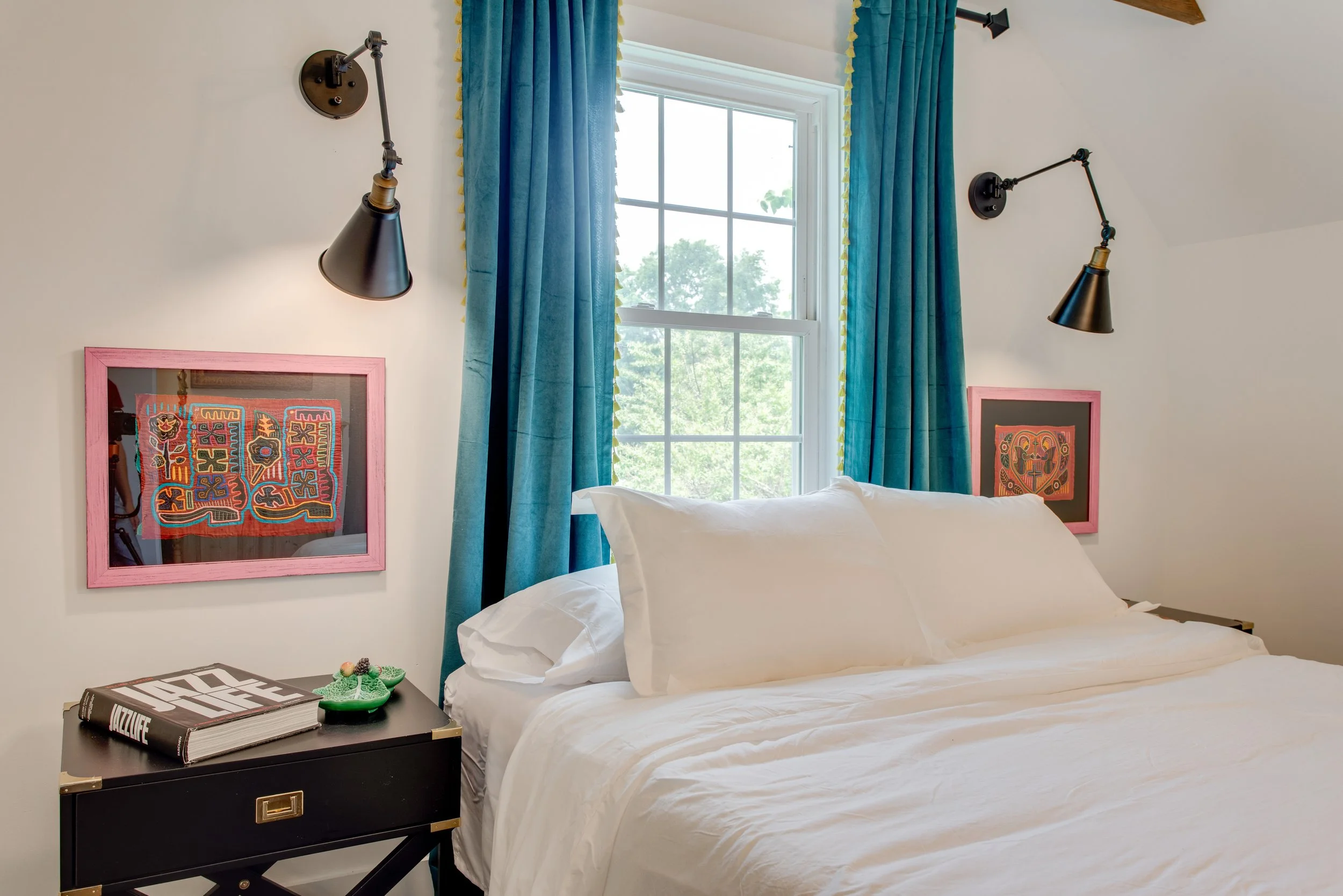 A bedroom with a bed covered in white sheets, a black nightstand with a book and green dinosaur toy, two black wall-mounted reading lamps, colorful framed artwork on the wall, and a window with blue curtains showing trees outside.