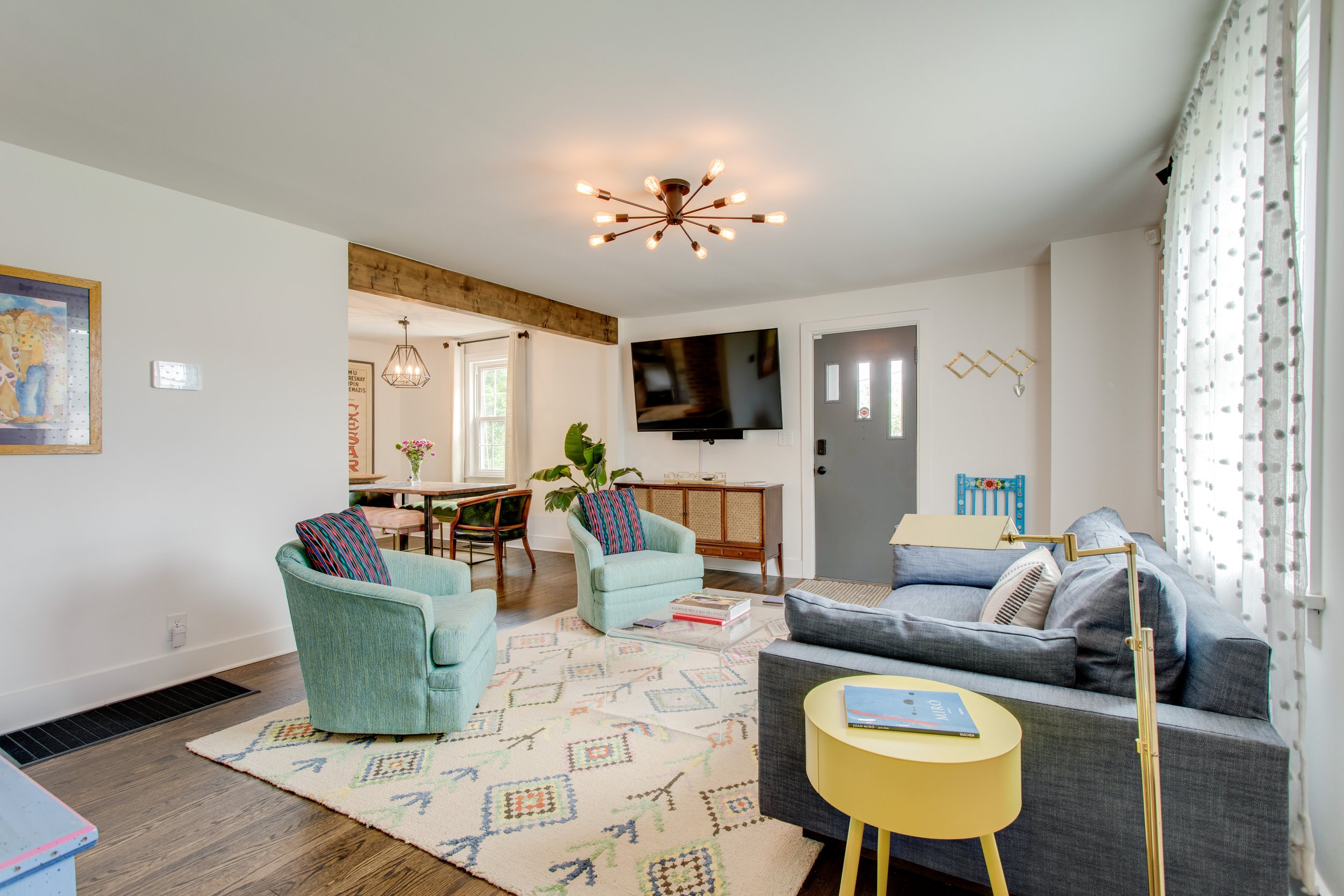 Living room with a gray sofa, two mint green armchairs, a yellow side table with a book, a large patterned area rug, a wall-mounted TV, a window with sheer curtains, and a modern chandelier on the ceiling.