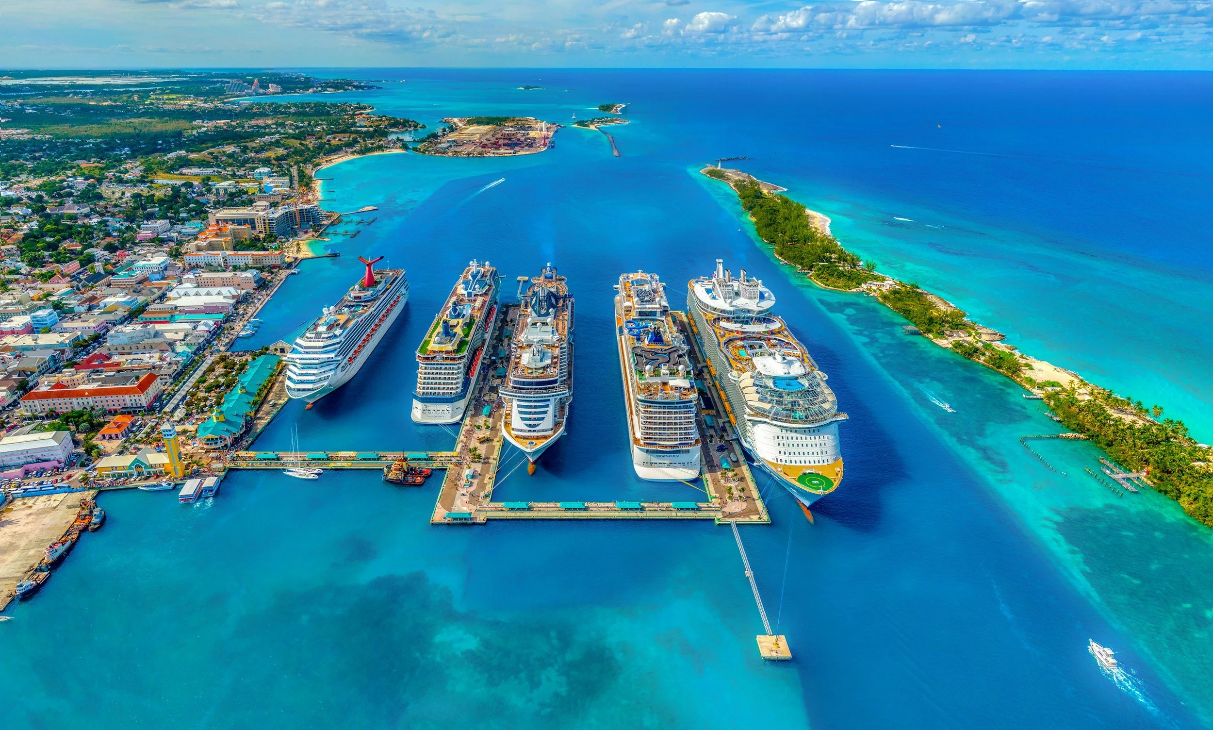 Aerial view of a cruise ship port in a tropical location with five large cruise ships docked, surrounded by turquoise water, a city with buildings and greenery on the left, and a narrow island with trees on the right.