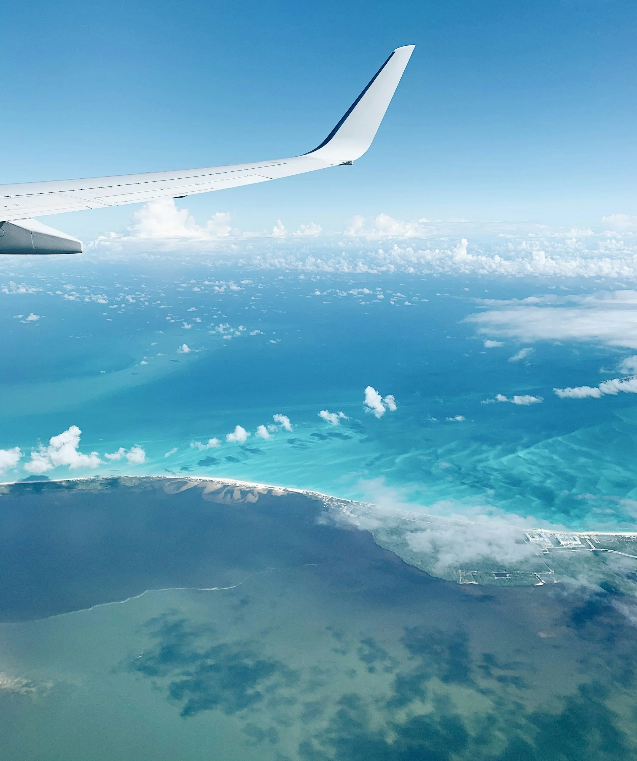 View from an airplane window showing the wingtip, turquoise waters, white clouds, and a coastline.
