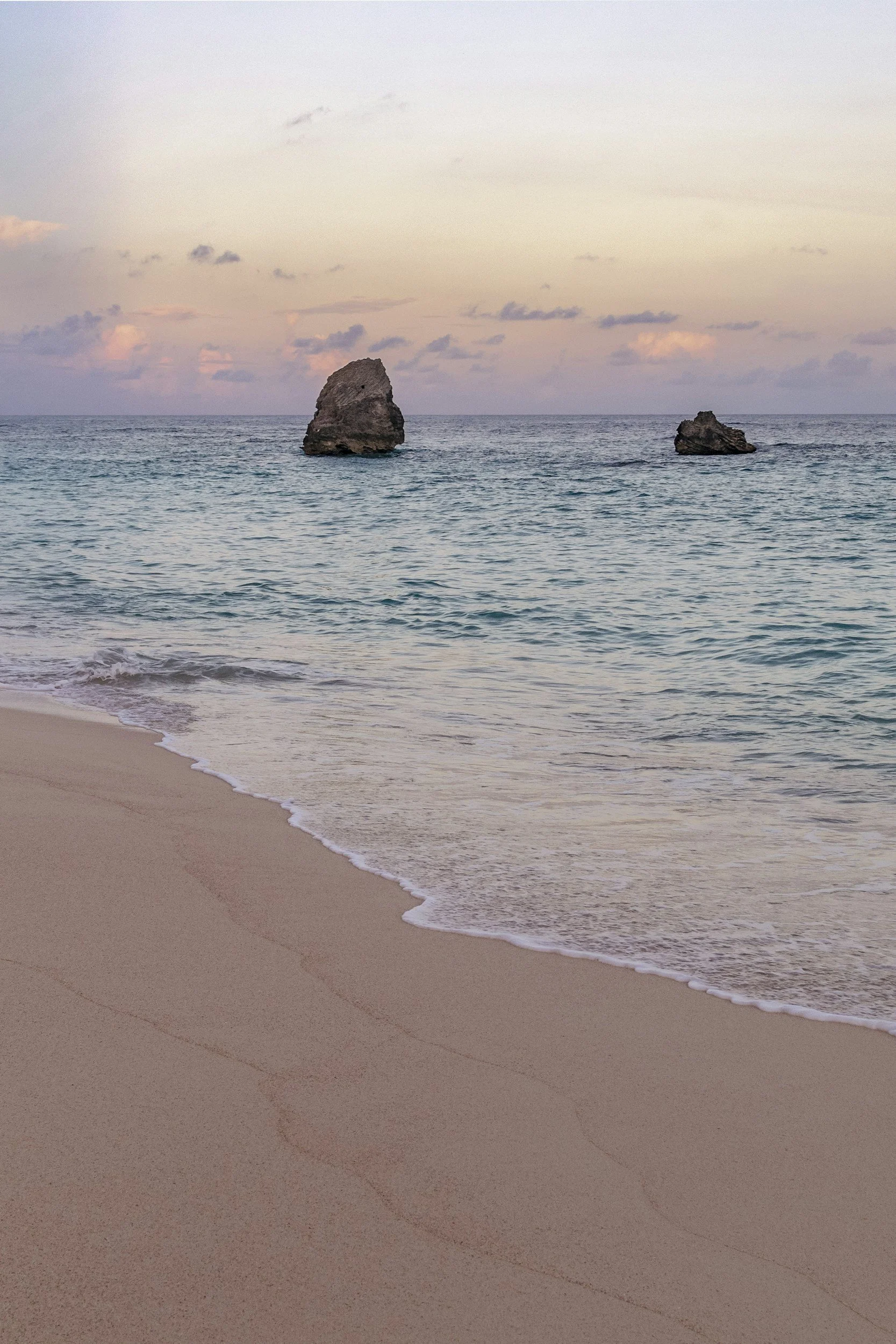 A beach scene with two large rocks in the ocean under a cloudy sky during sunset or sunrise.