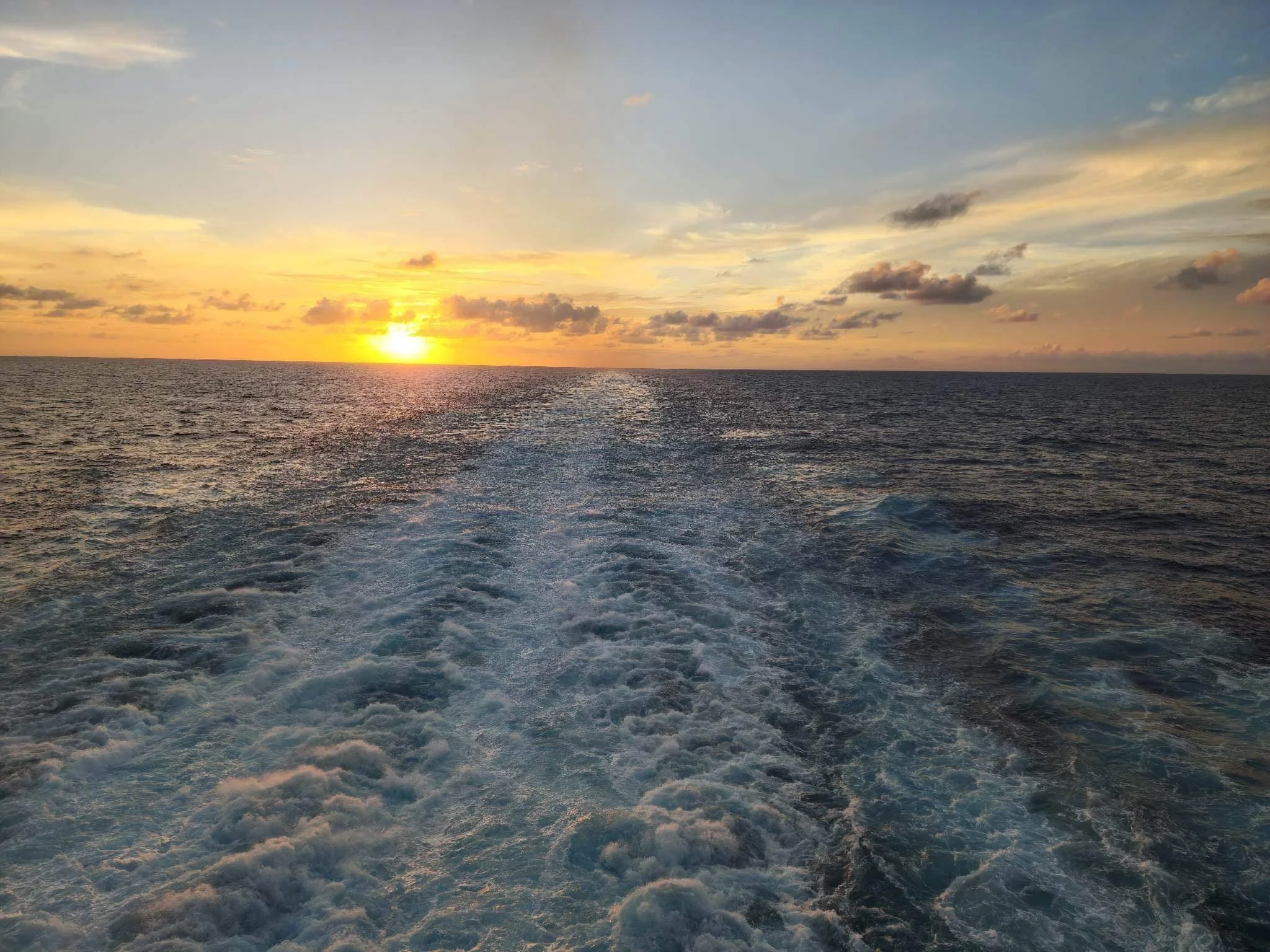 Sunset over the ocean with clouds and gentle waves, as seen from a boat with a wake in the water