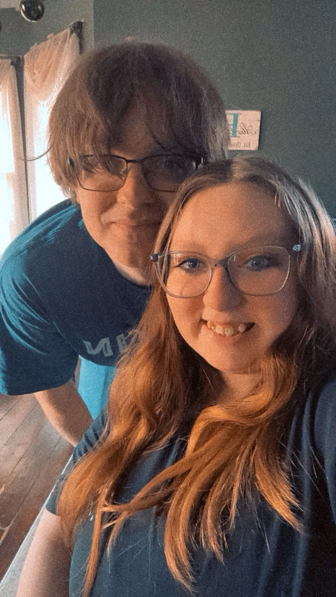 A young woman with long red hair and glasses smiling, with a young man with wavy hair and glasses leaning over her from behind, in an indoor setting with wood flooring and a dark green wall.