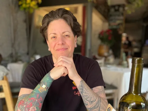 A woman with short dark hair and tattoos on her arm, sitting at a table outdoors. She is smiling with her hands clasped under her chin and wearing a black t-shirt. In the background, there is a blurred outdoor setting with plants and table decorations.