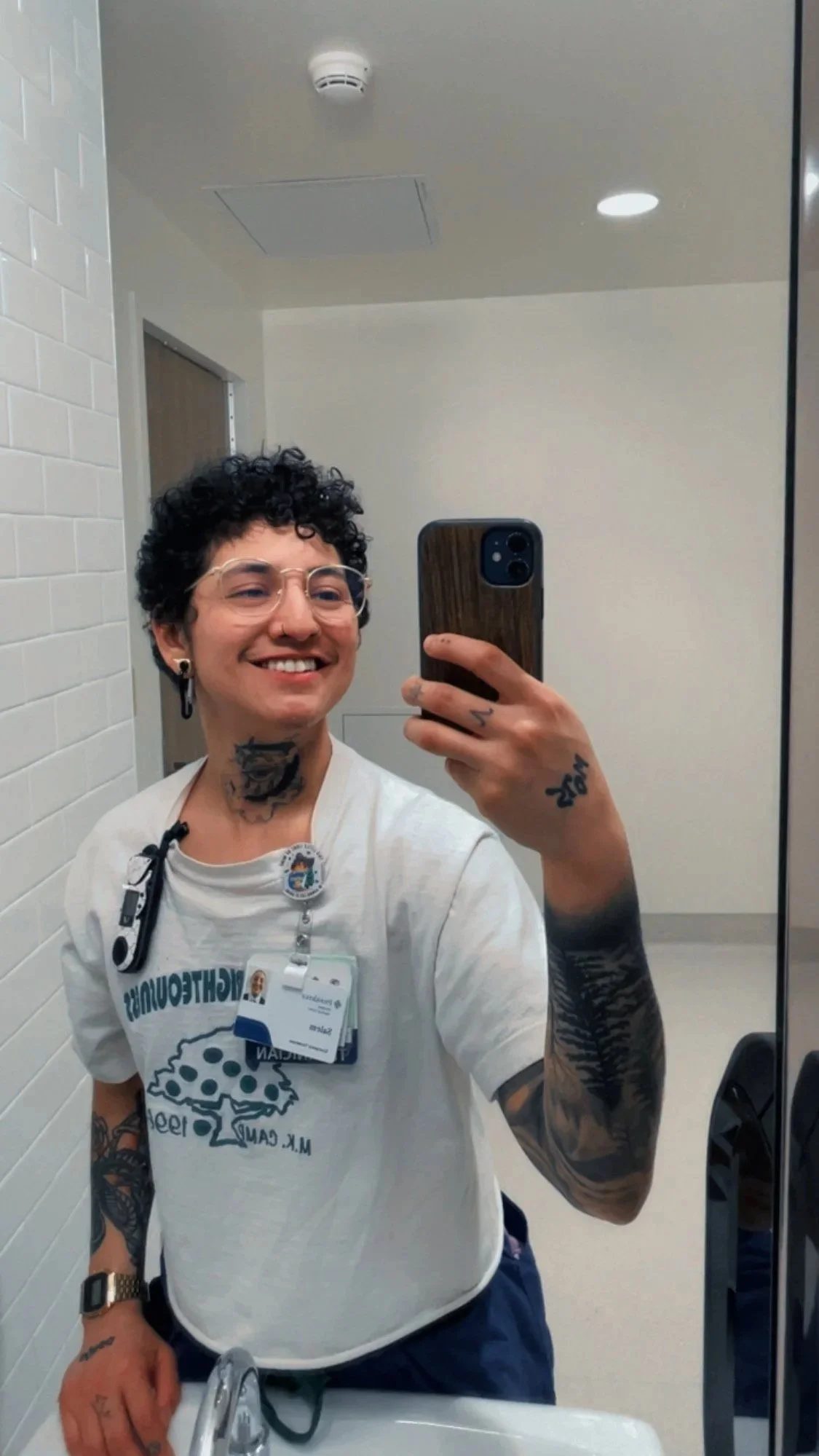 A man with short curly black hair and glasses taking a mirror selfie in a room, smiling, wearing a white t-shirt with a logo, and has tattoos on his neck, arms, and hand.