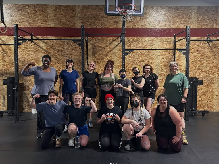 Group of eleven people posing in a gym with workout equipment and basketball hoop in the background.