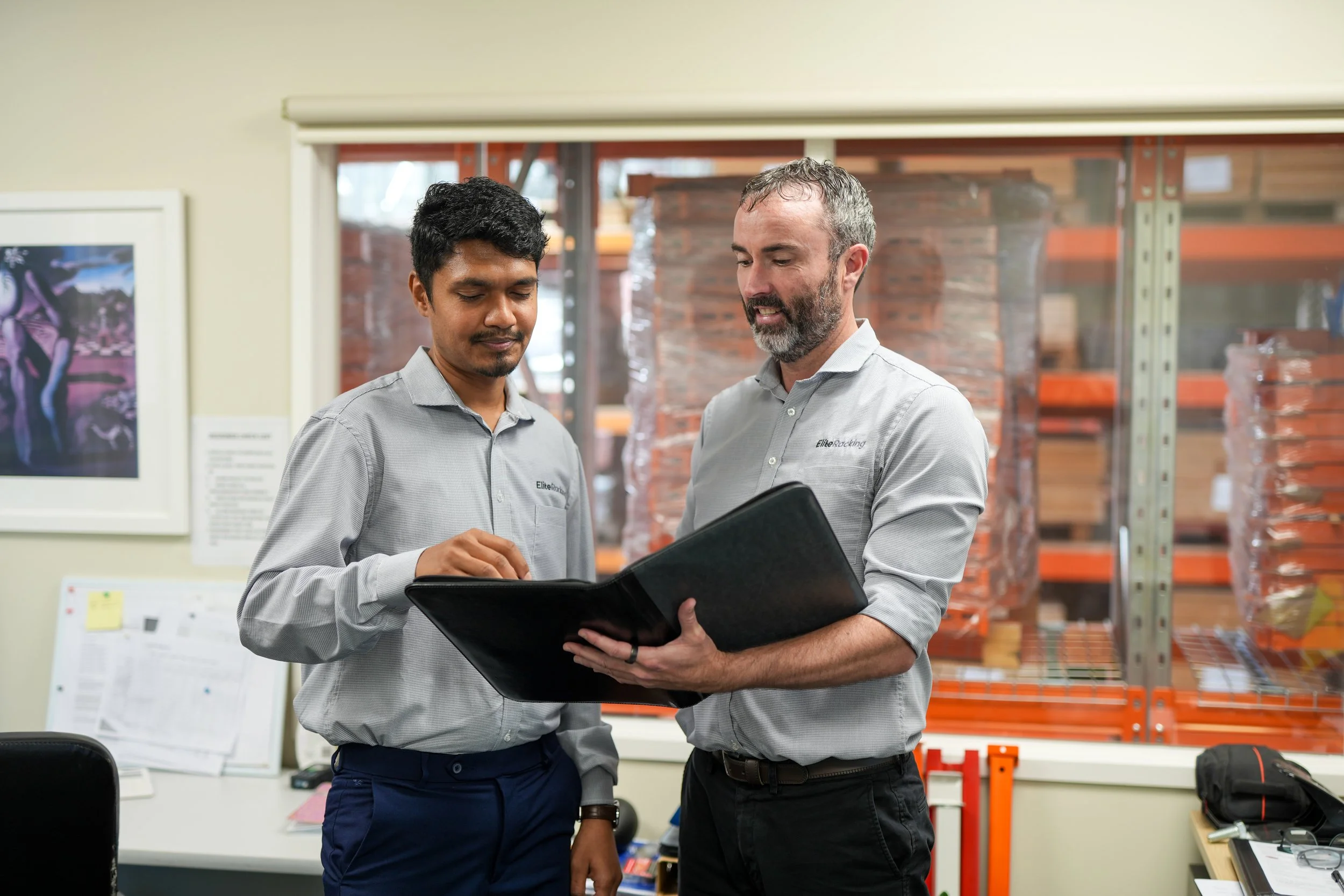 Two men in work shirts reviewing documents in an office or warehouse.