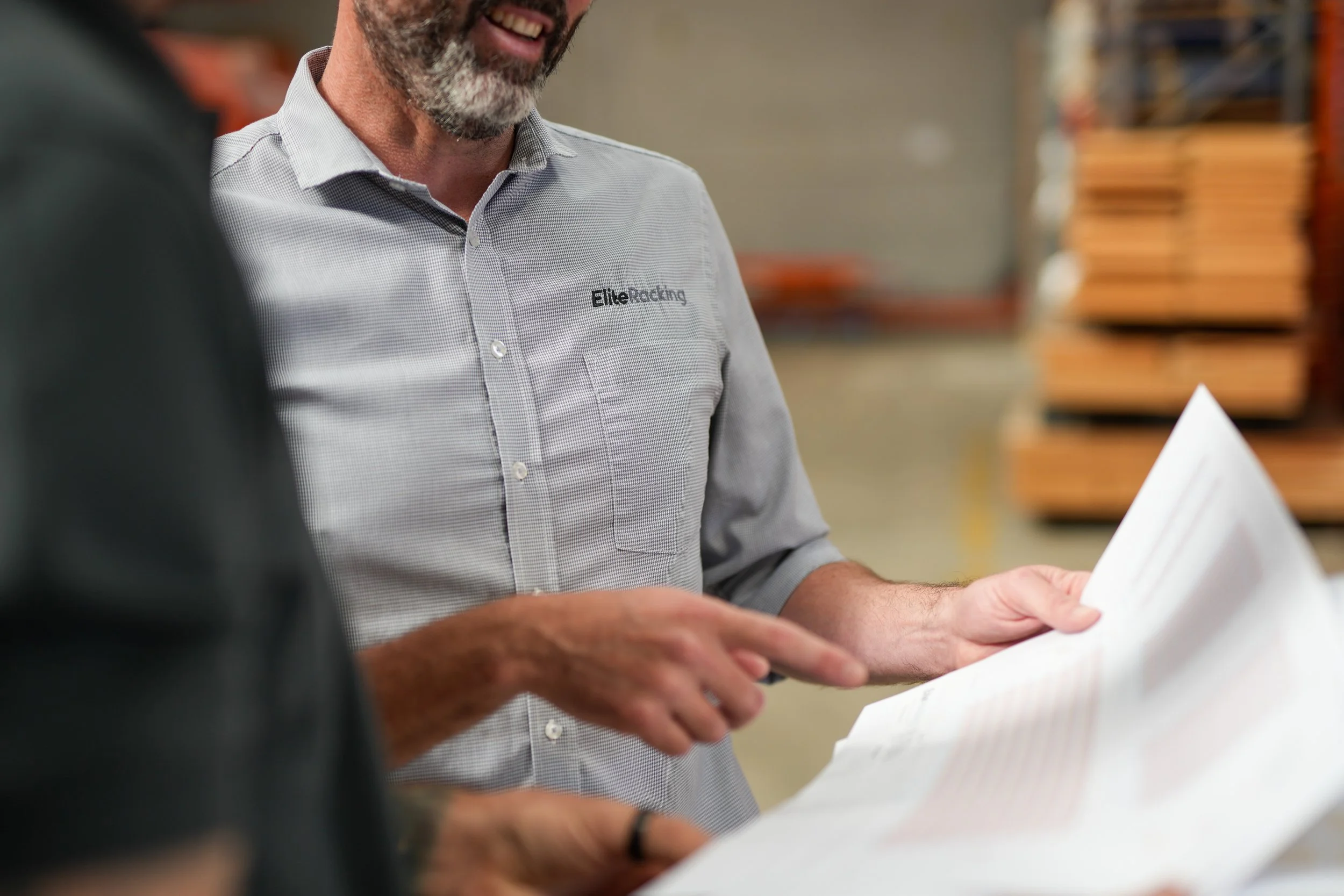 A man in a gray work shirt with the logo 'Elite Racking' explaining plans to another person in a warehouse.