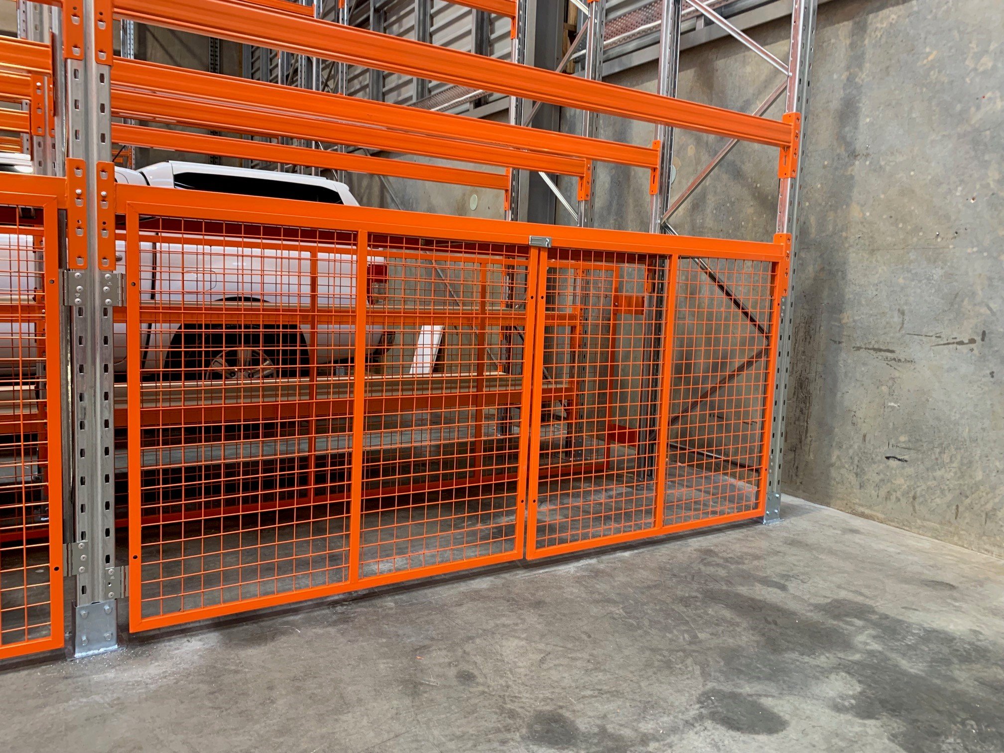 Orange safety gate blocking access to stacked metal shelving in a warehouse with a white vehicle visible behind the gate.