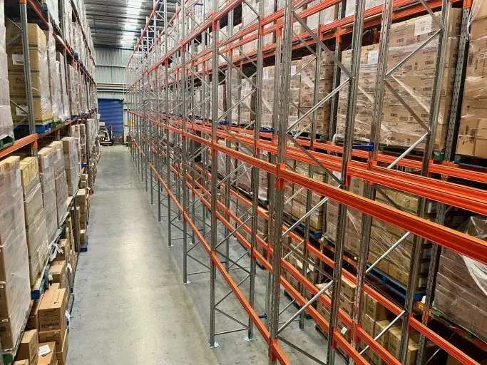 Empty industrial warehouse aisle with tall metal and orange shelving, some shelves partially filled with boxes.