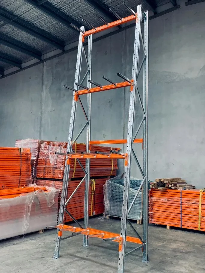 A tall metal shelving unit with orange beams and gray vertical supports, standing in a warehouse with stacks of orange and gray metal pipes and other materials in the background.