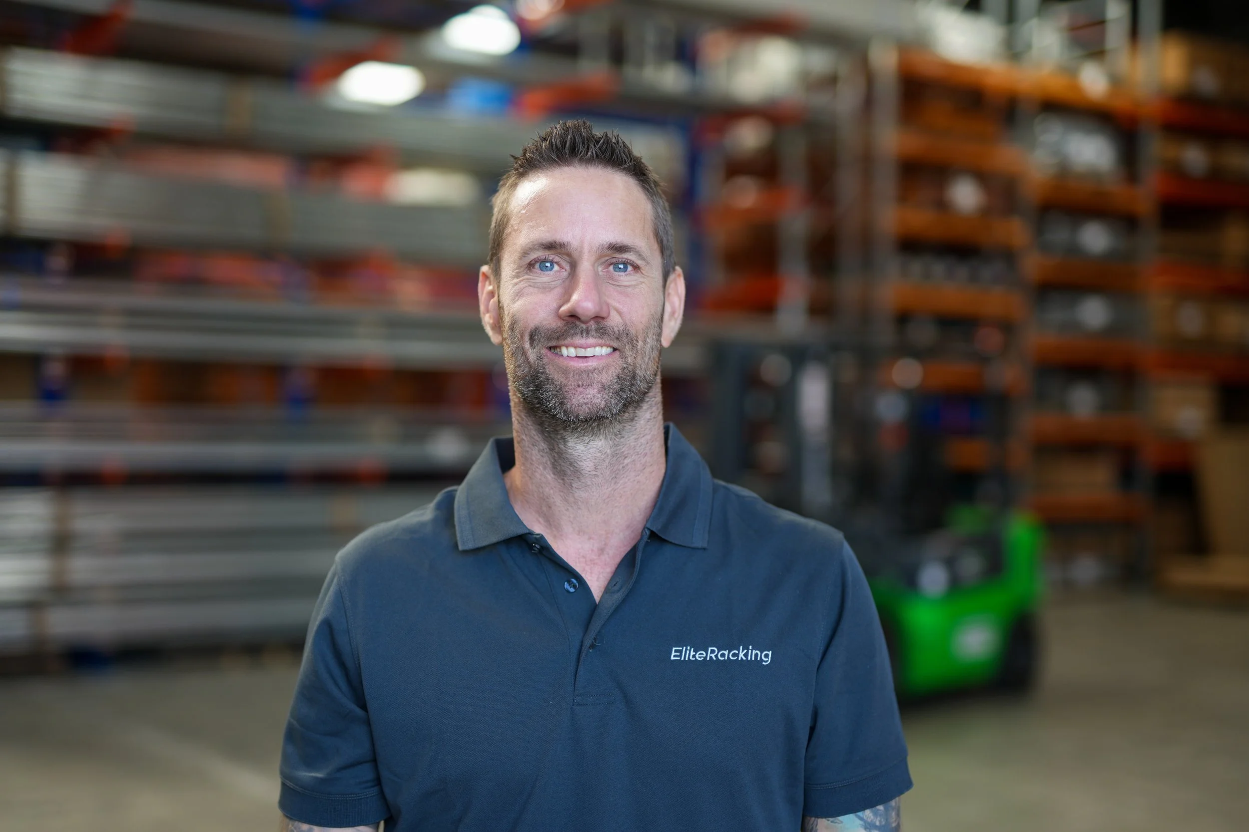 A man with short brown hair, blue eyes, and a beard, smiling, standing in front of industrial shelving with metal and wooden pallets in a warehouse. He is wearing a navy blue polo shirt with 'EliteRacking' on it.