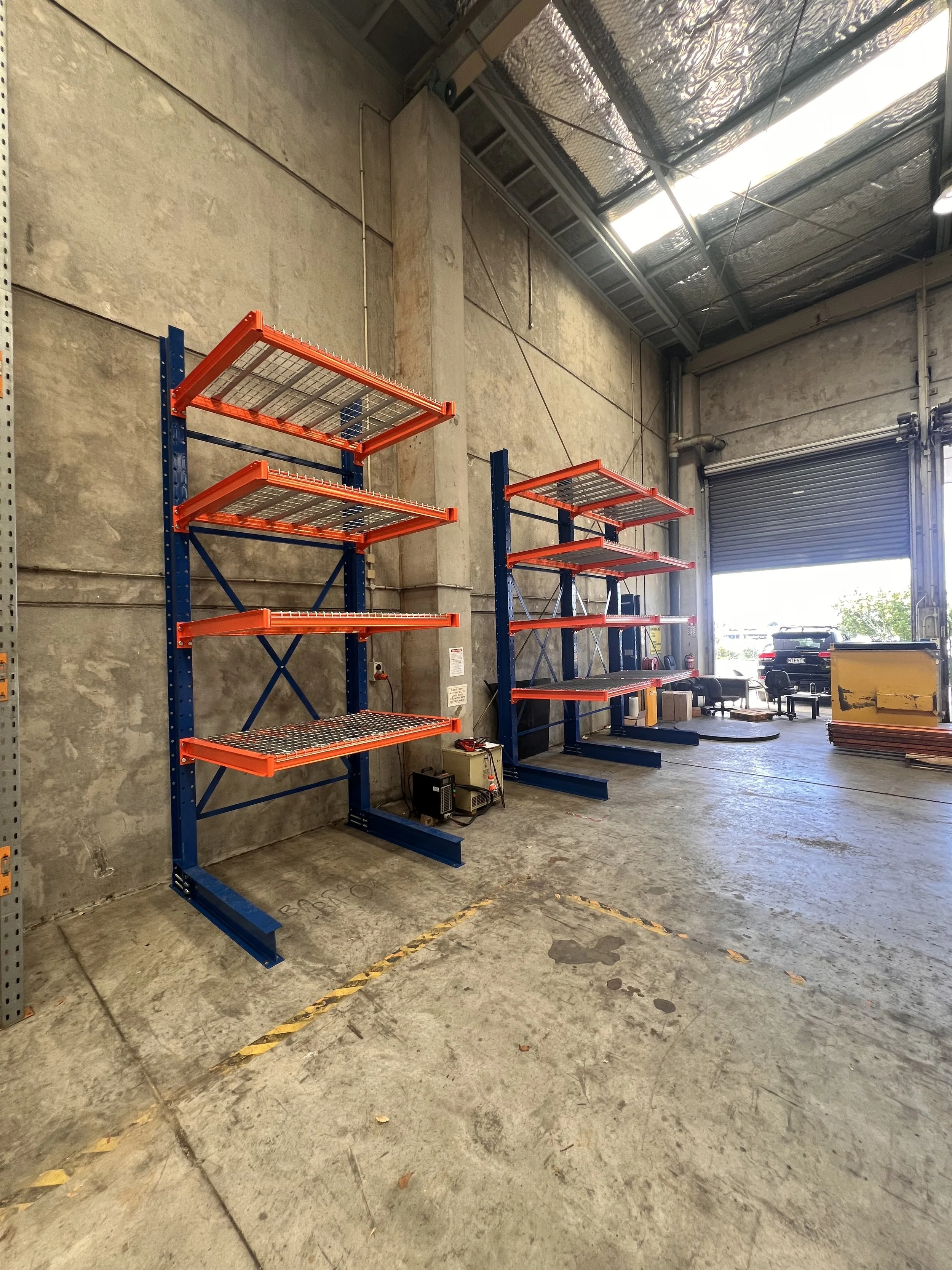 Empty industrial warehouse with two sets of blue and orange metal shelving units near a large open garage door, with some assorted items and vehicles outside.