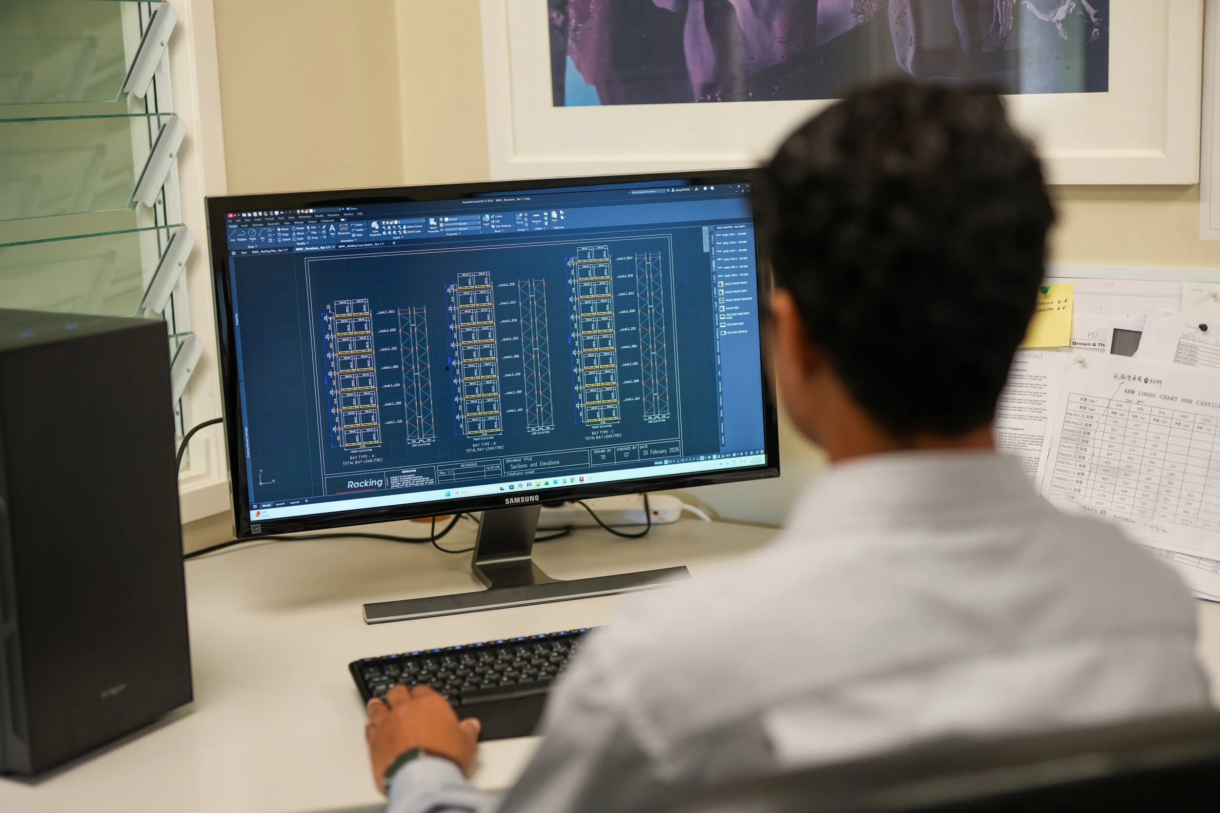 A person working at a computer with engineering drawings on the screen and papers on the desk. The drawings appear to be technical schematics or blueprints.
