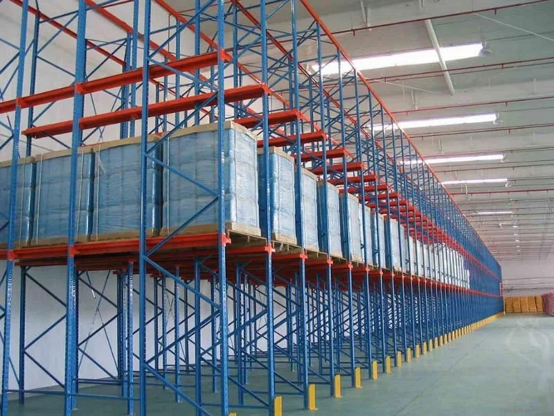 Warehouse with tall blue and orange metal shelving units holding pallets of goods wrapped in plastic.
