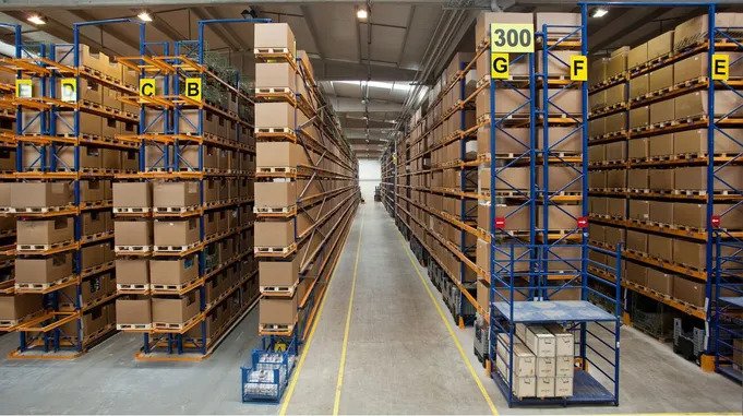A large warehouse aisle with tall metal racks filled with cardboard boxes and containers, organized and labeled with letters and numbers.