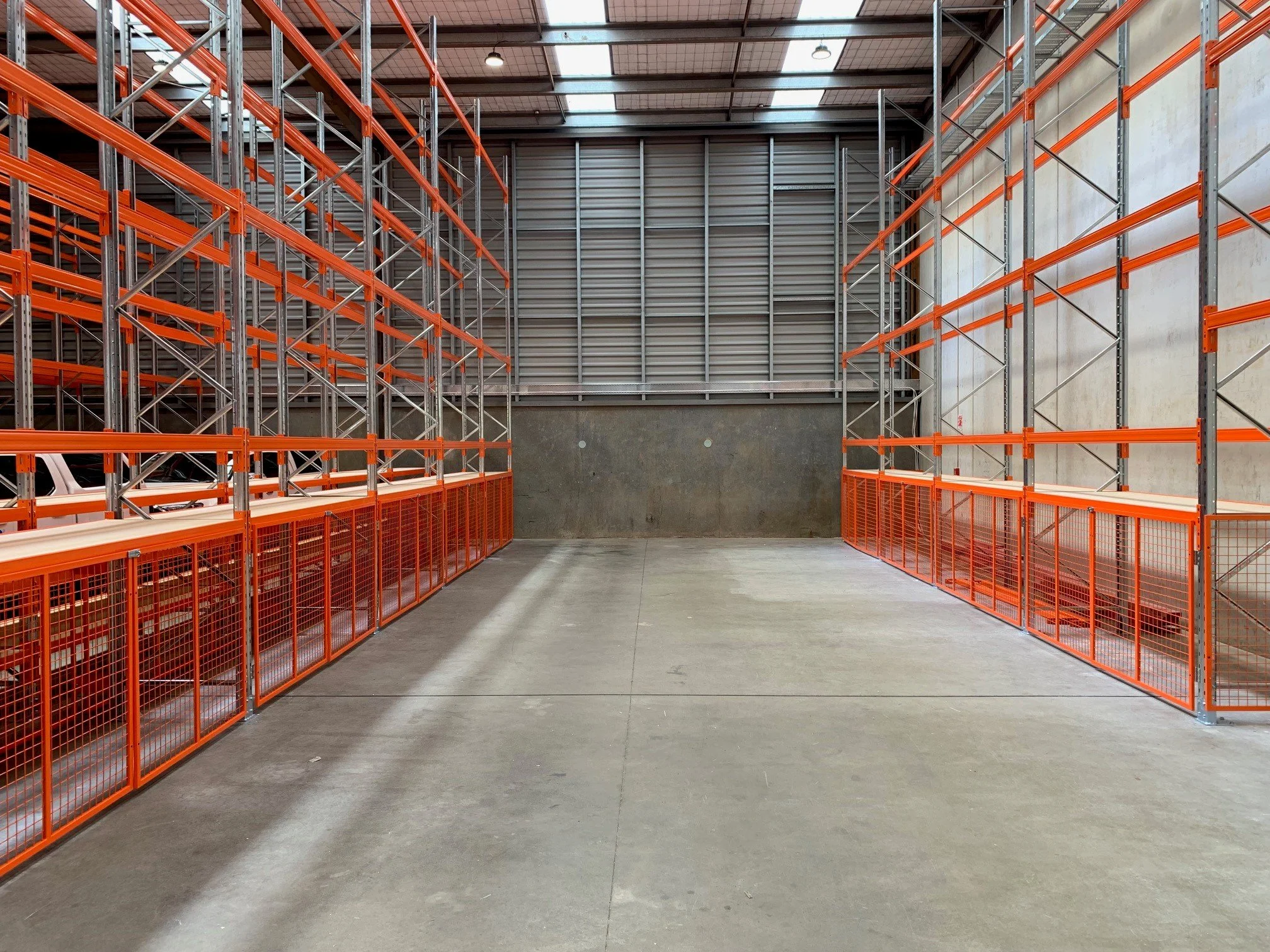 Empty warehouse storage aisle with orange metal shelving units on both sides, concrete floor, and metal wall panels.