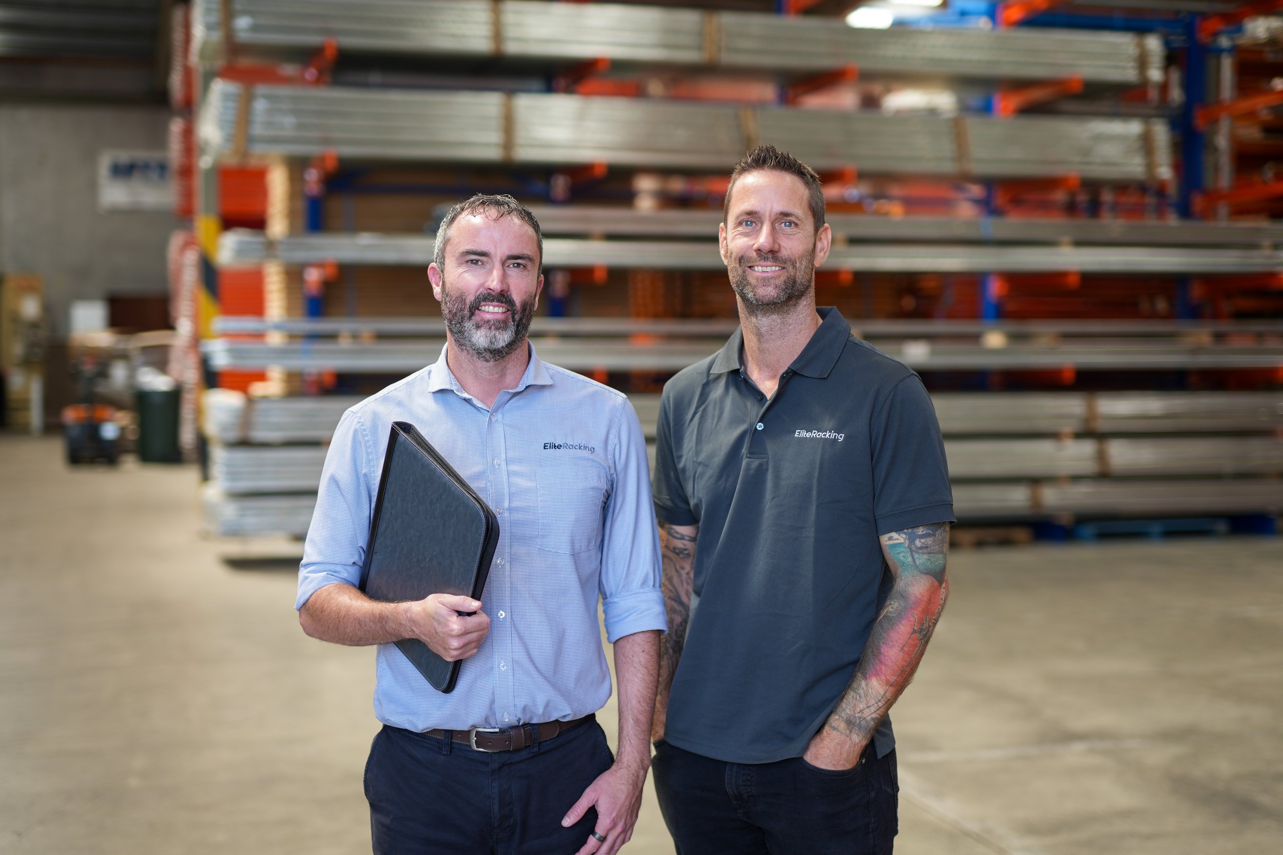 Two men standing inside a warehouse with shelves of metal and construction materials in the background. One is holding a black folder, and both are smiling. They are wearing collared shirts with the logo 'EliteRacking'.