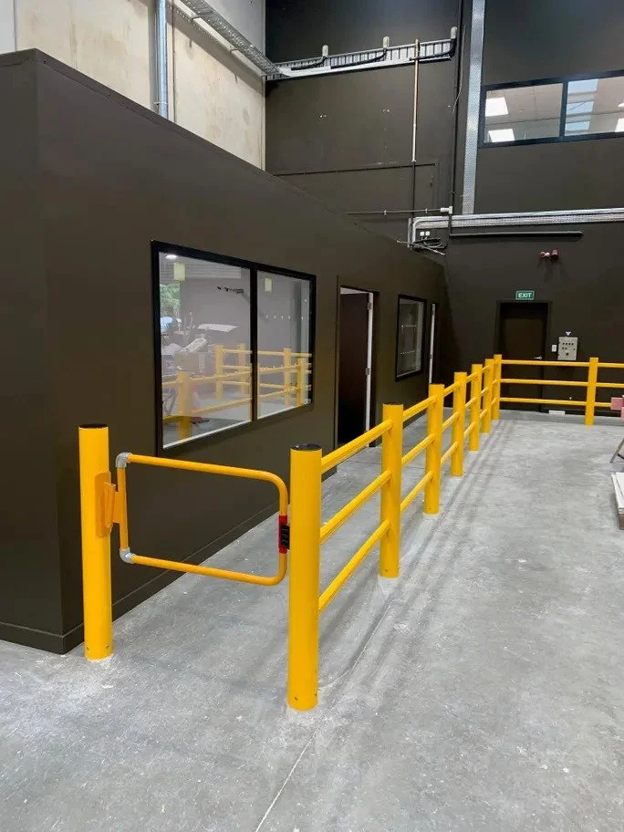 A yellow safety railing and gate in front of a dark-colored building with windows and a door, in an industrial or warehouse setting.