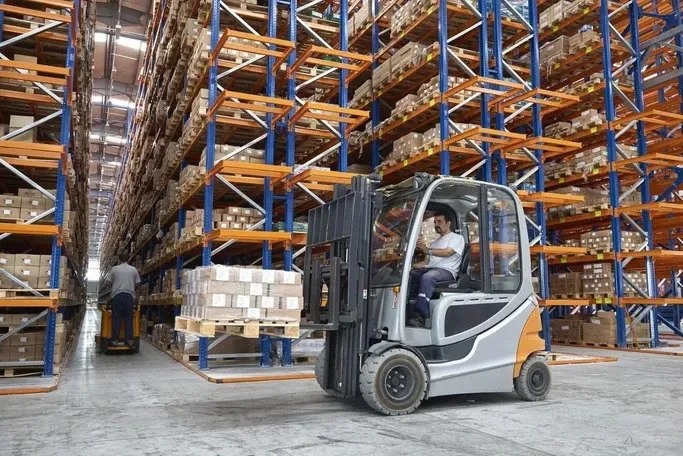 A person operating a forklift in a large warehouse filled with tall shelves stocked with boxes and pallets.