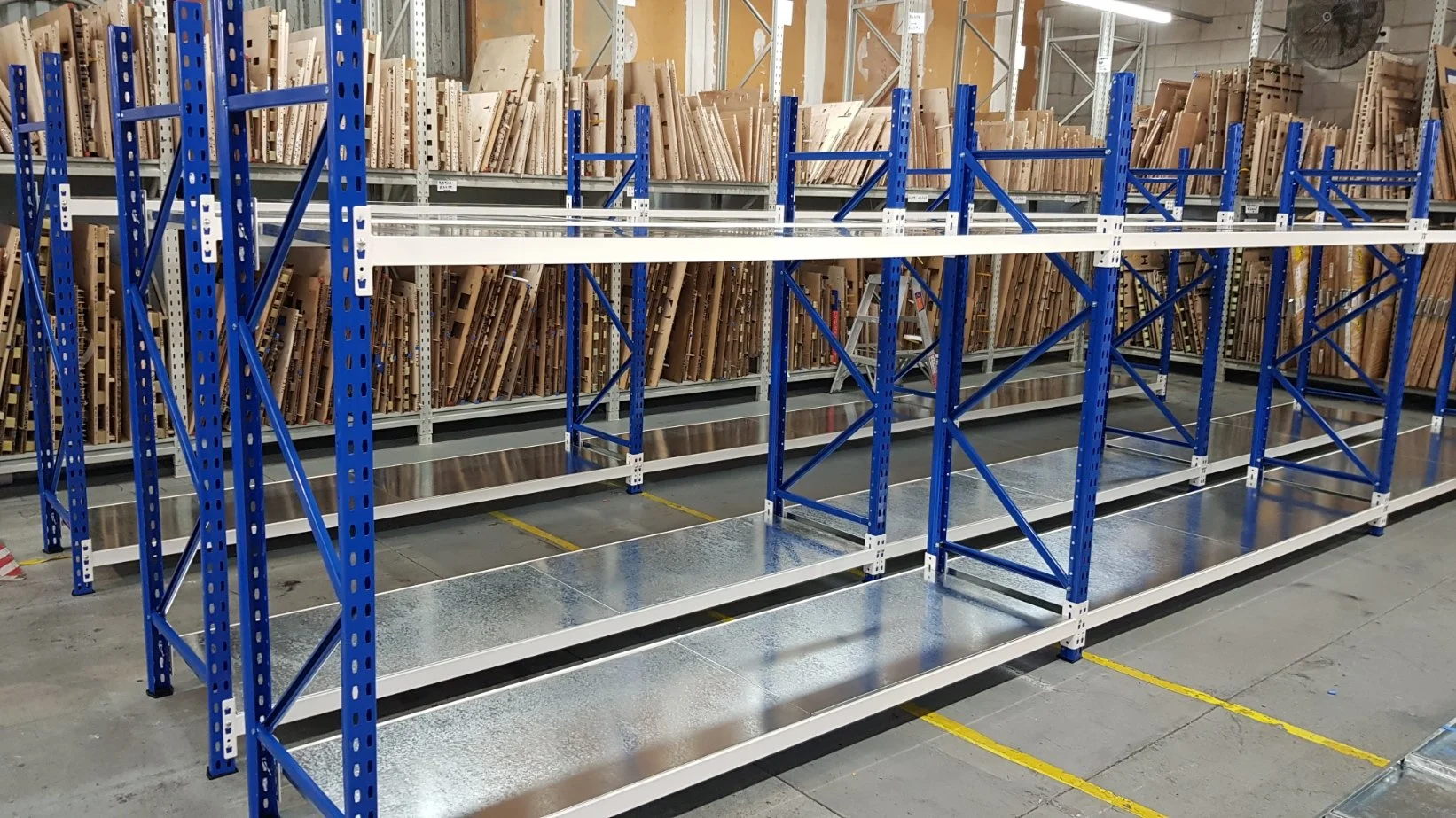 Empty blue and white metal shelving units in a warehouse, with some wooden boards stored on high shelves in the background.