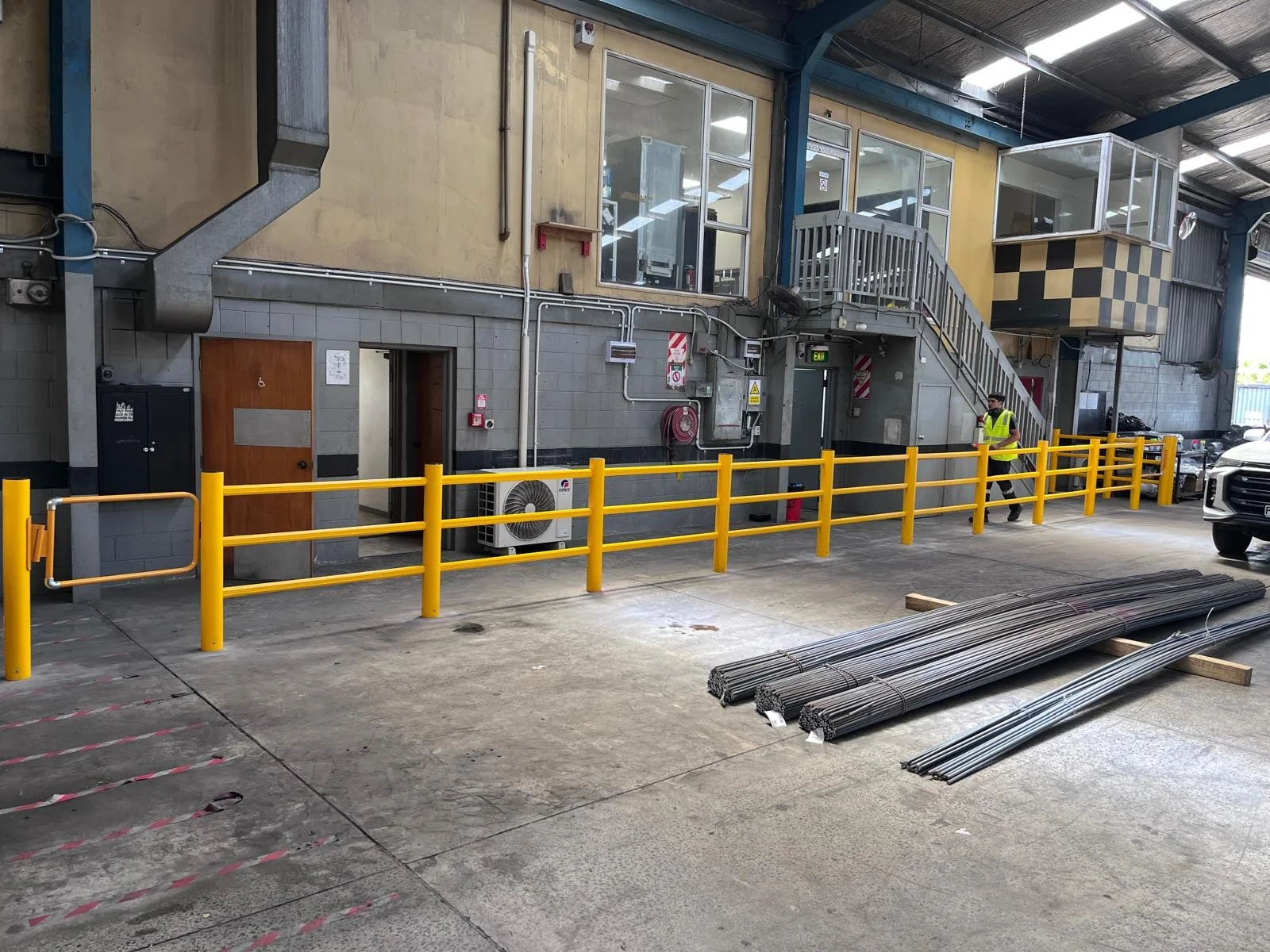 Indoor industrial warehouse with yellow safety barriers, metal pipes on the ground, a worker in a yellow safety vest, and parked vehicles.