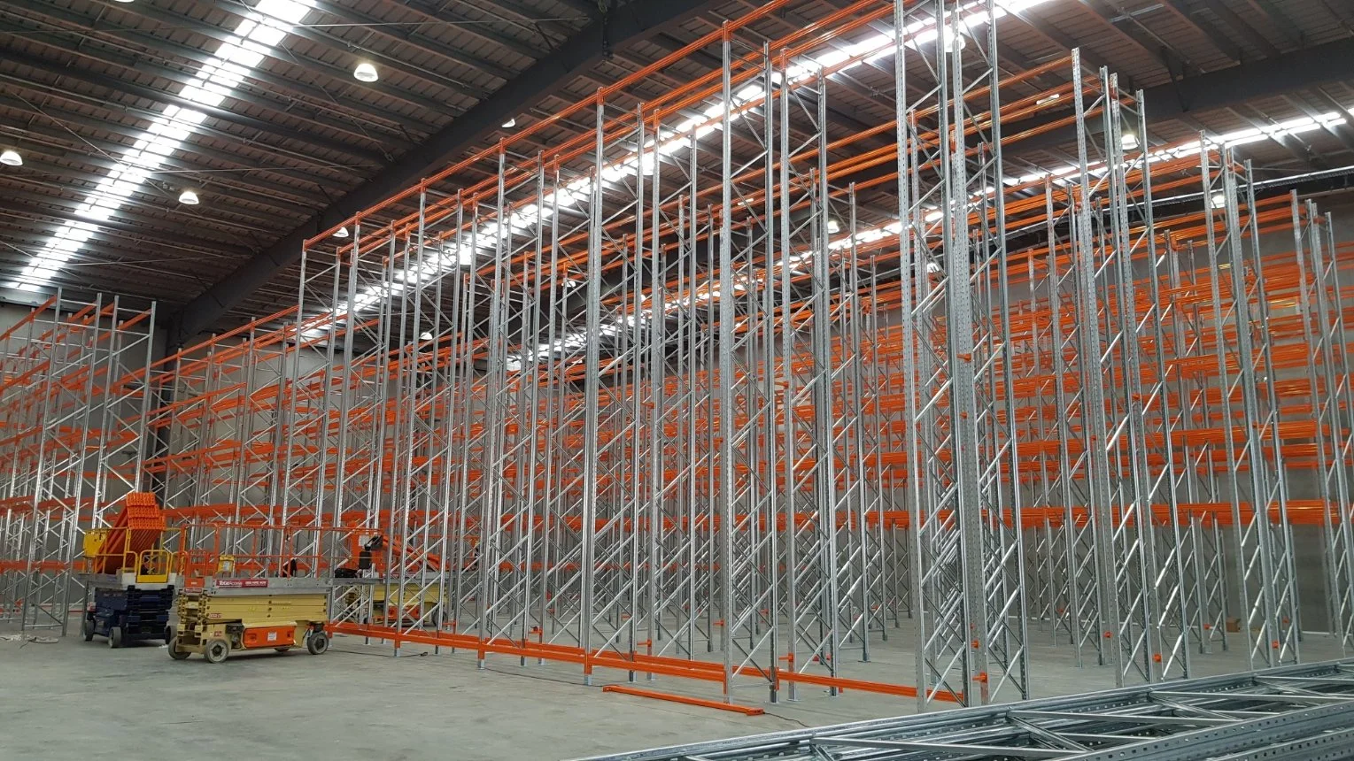 Inside a large warehouse with metal racks under construction, with orange beams and tools on the ground.
