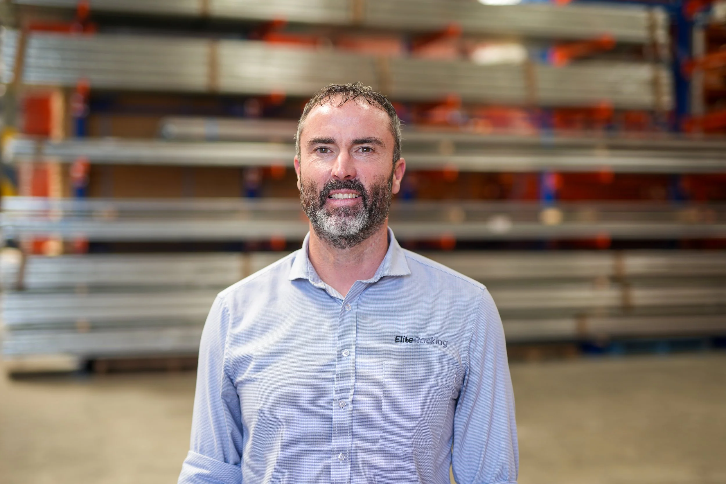 A man with a beard and short hair, wearing a light blue button-up shirt with the logo 'Elite Racking,' standing in front of industrial shelving units stored with metal beams in a warehouse.