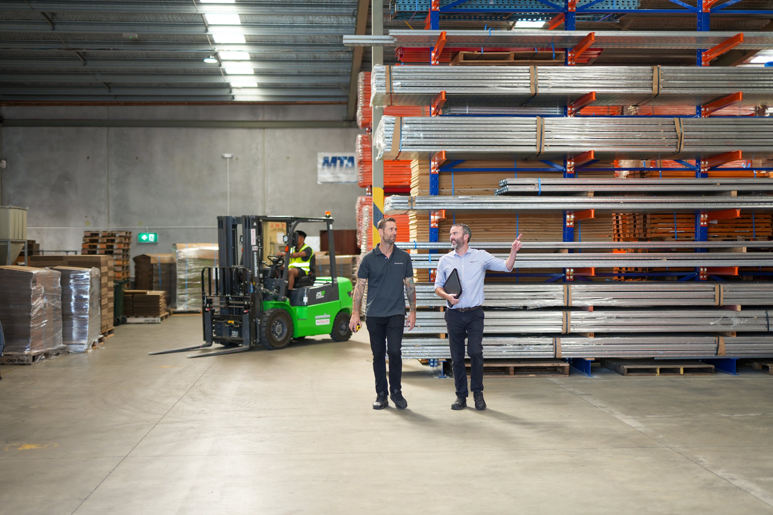 Two men walking and talking in a warehouse with stacked metal pipes and a forklift moving in the background.
