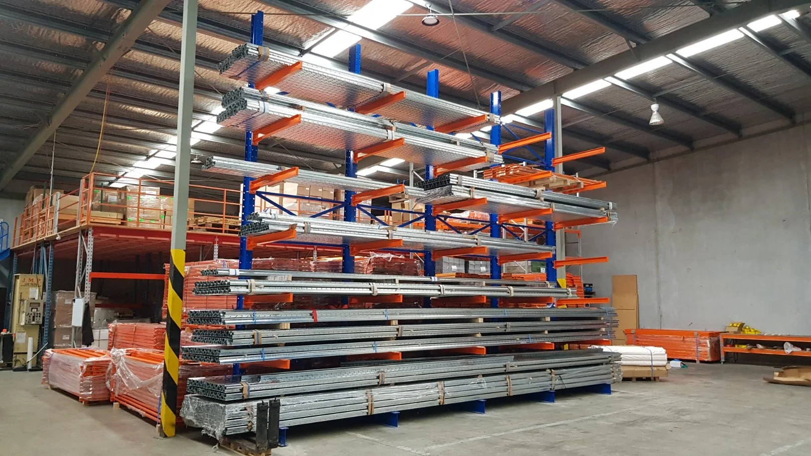 Industrial storage warehouse with metal scaffolding, pipes, and materials stacked on shelves and on the floor.