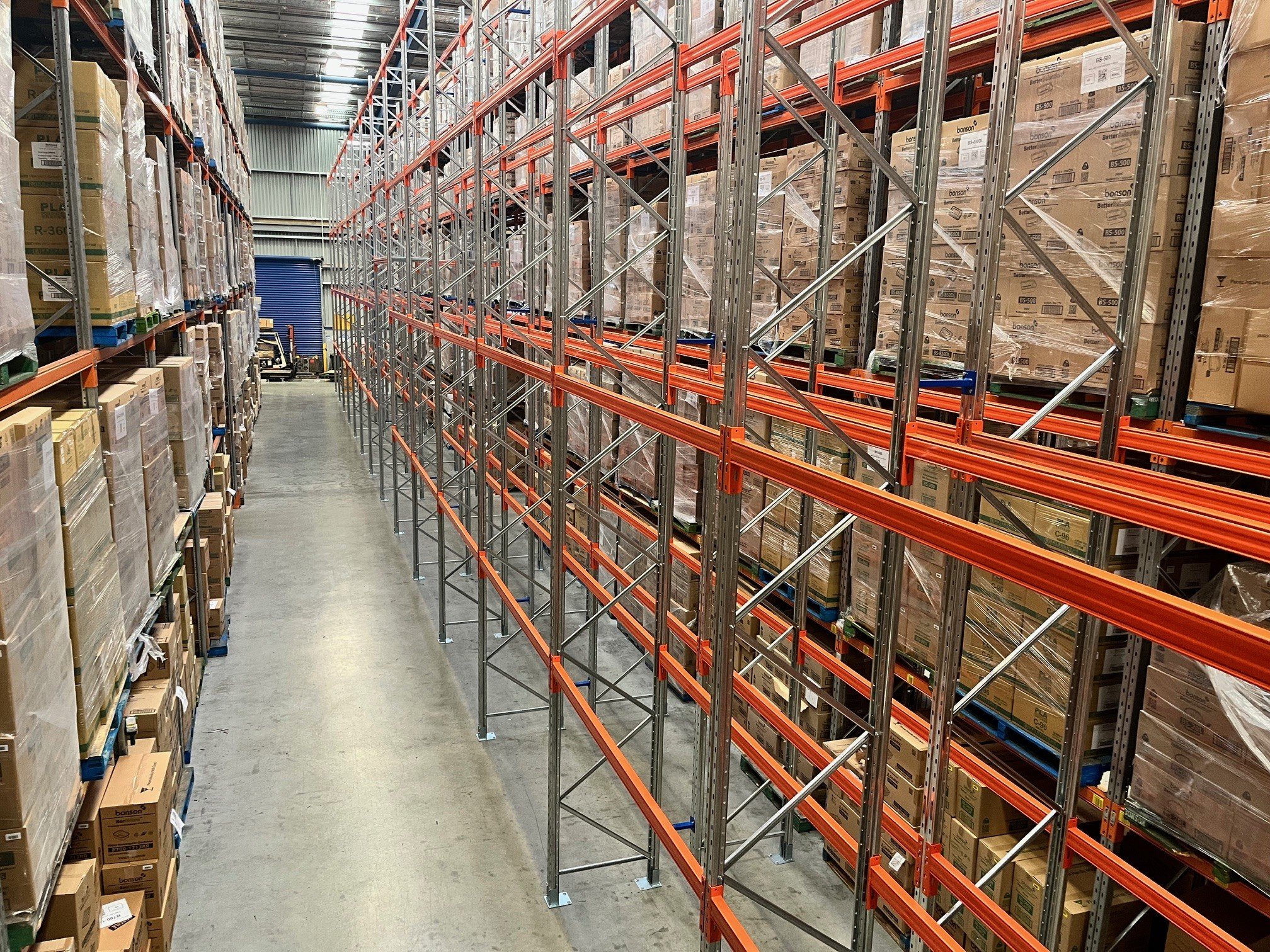 Empty store warehouse aisle with tall shelves stocked with boxes and pallets, metal racks on the right side, and a blue roller door in the background.