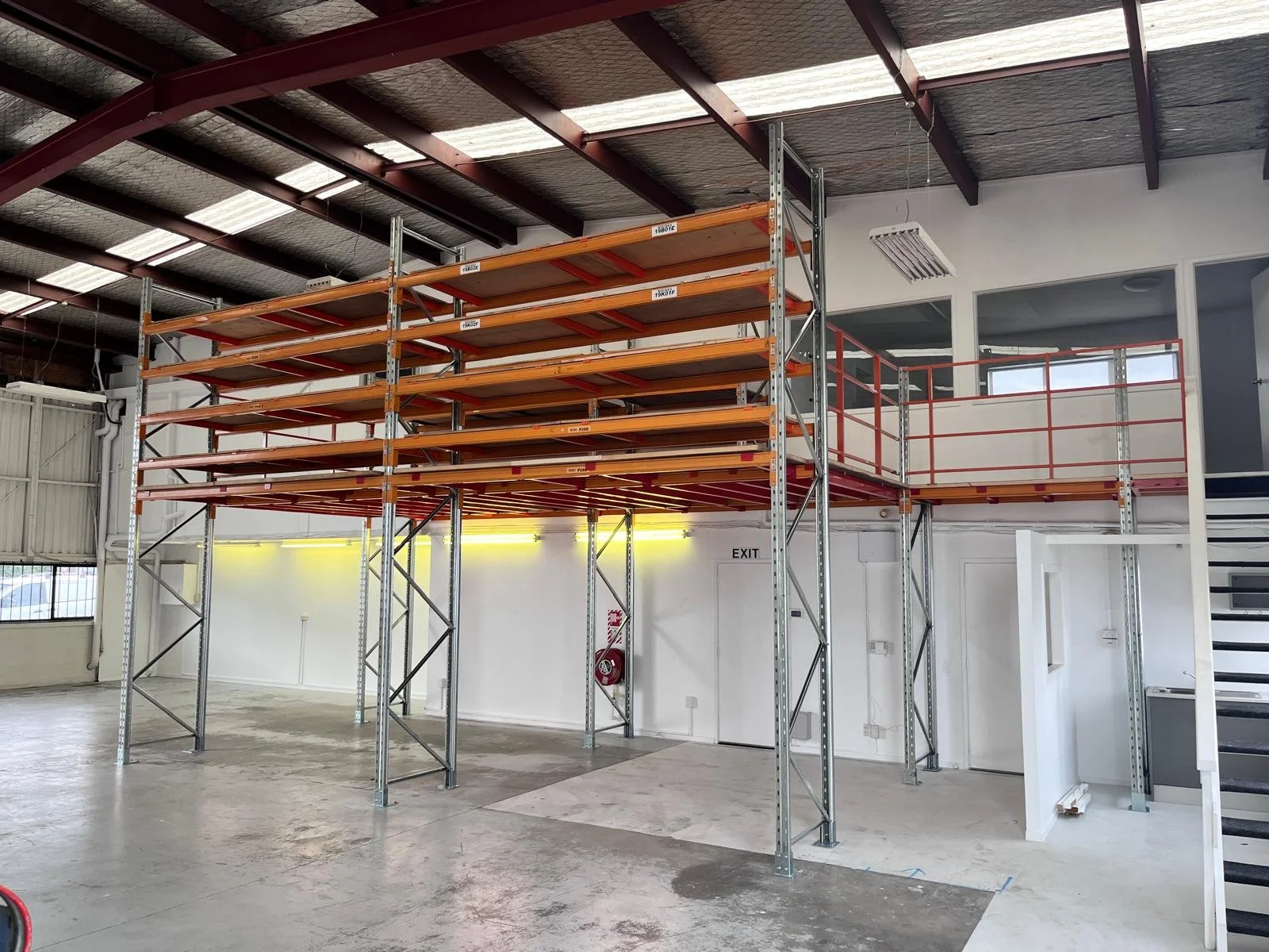 Empty industrial warehouse with metal storage racks and a staircase leading to a second level with a railing.