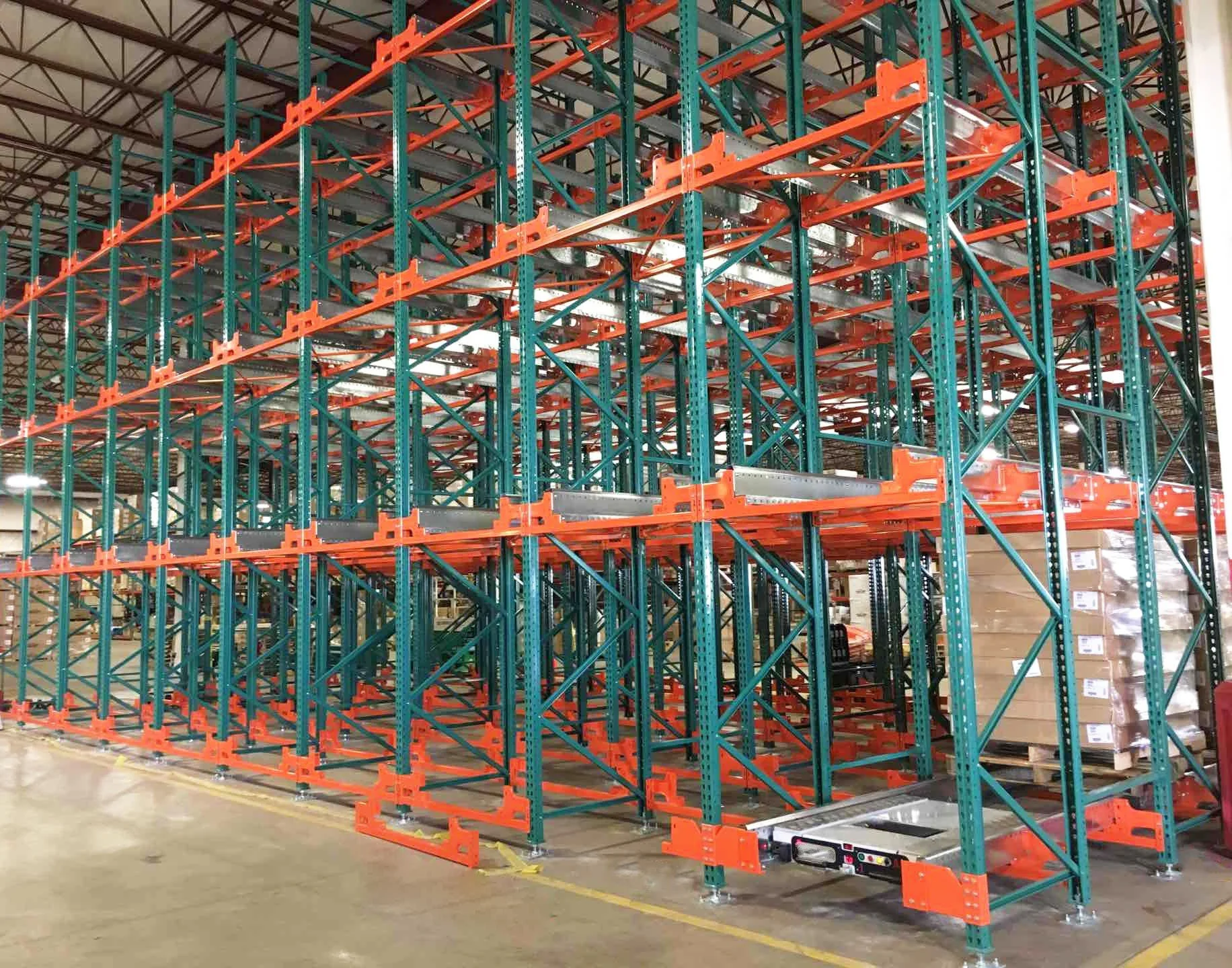 Industrial warehouse shelving unit with green and orange metal racks, partially loaded with boxes on pallets.
