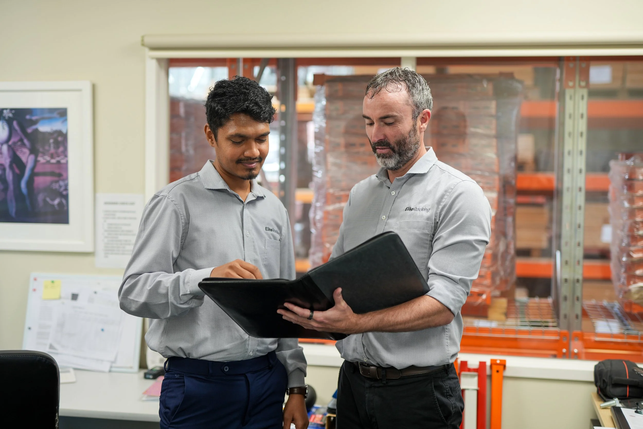 Two men in gray shirts reviewing documents together in a warehouse office.