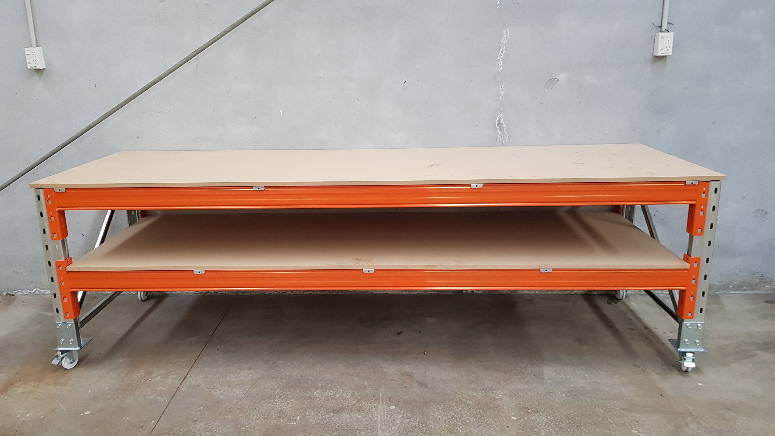 A two-tier industrial storage cart with beige wooden shelves and orange metal frame on caster wheels, positioned against a plain gray wall in a warehouse or workshop setting.