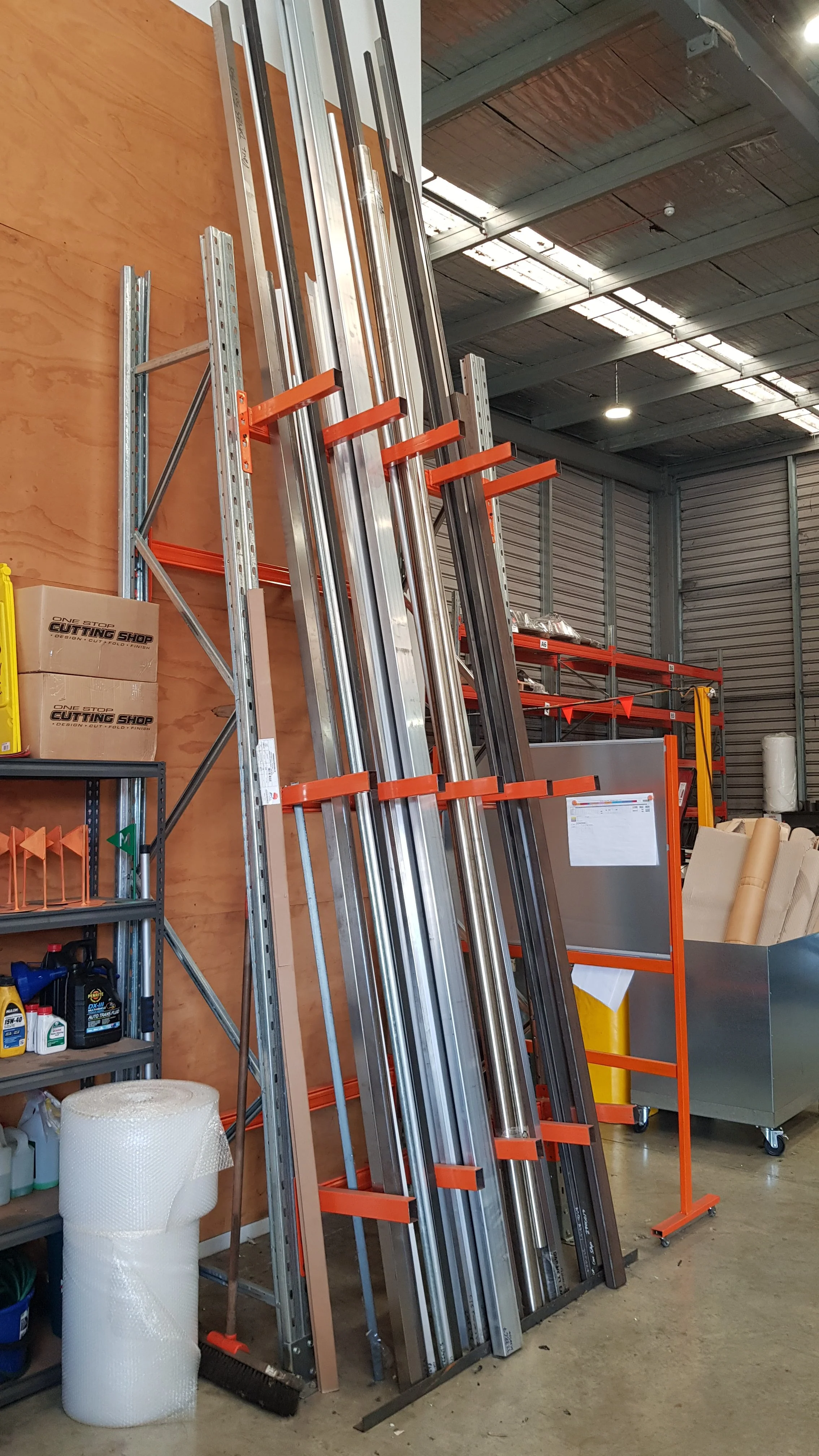 Metal shelving unit holding various long metal bars and pipes, with additional shelves containing boxes, cleaning supplies, and bubble wrap, in a warehouse setting.