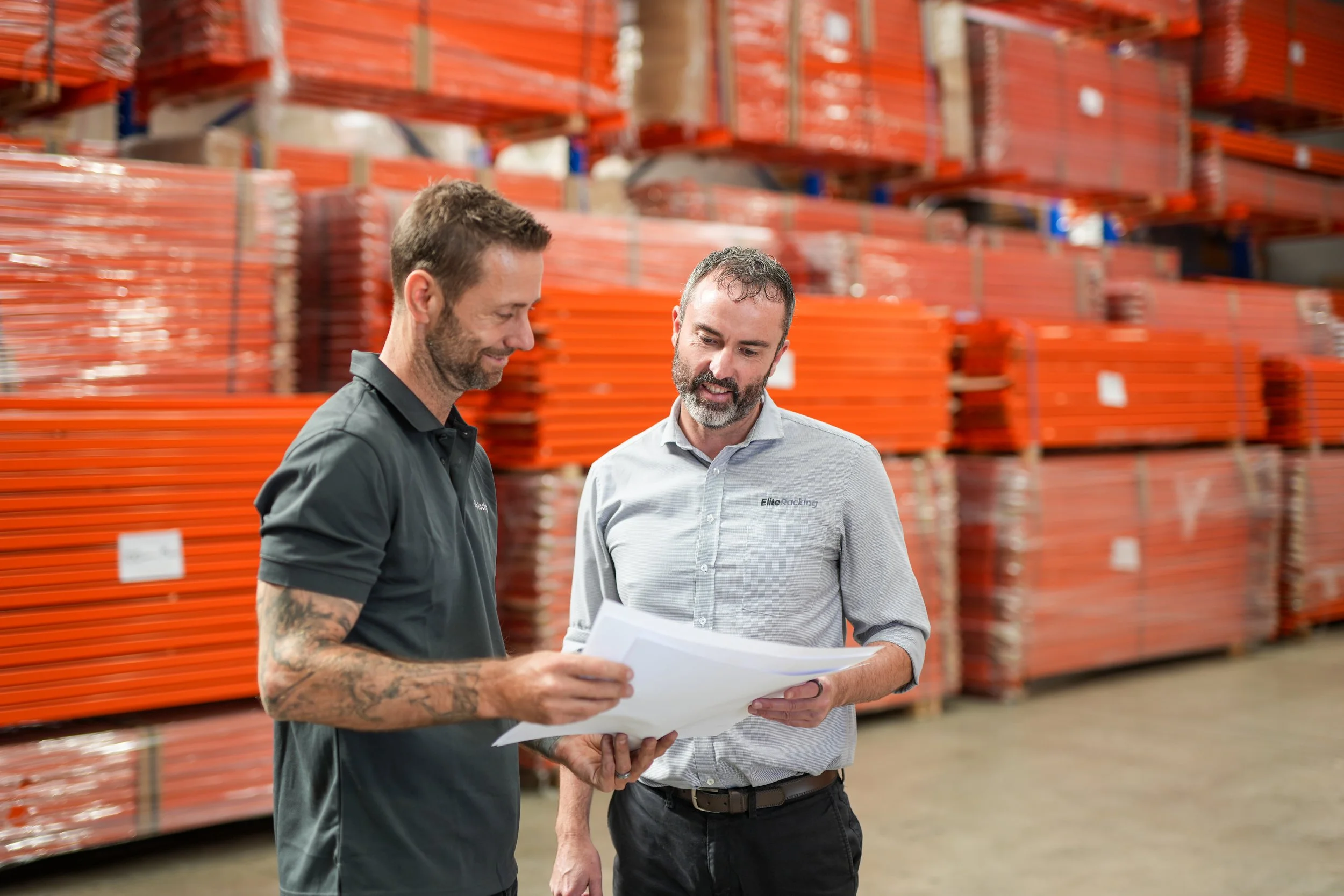 Two men in warehouse discussing plans or documents, with shelves of orange loading ramps or platforms in the background.