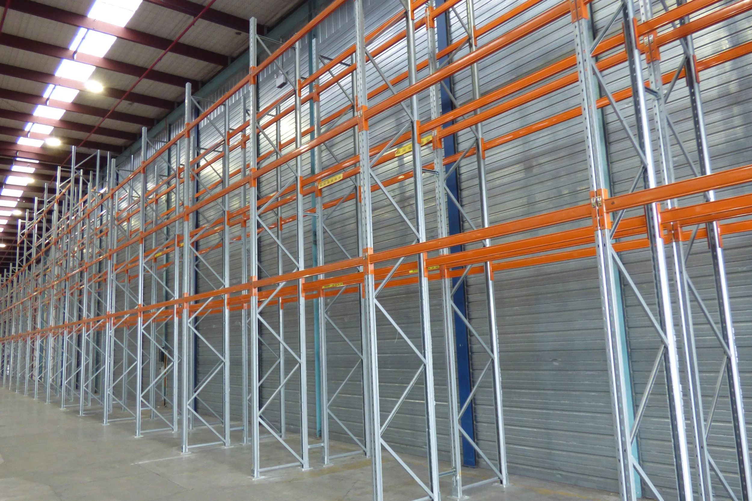 Empty industrial warehouse with tall metal racks and orange safety beams.