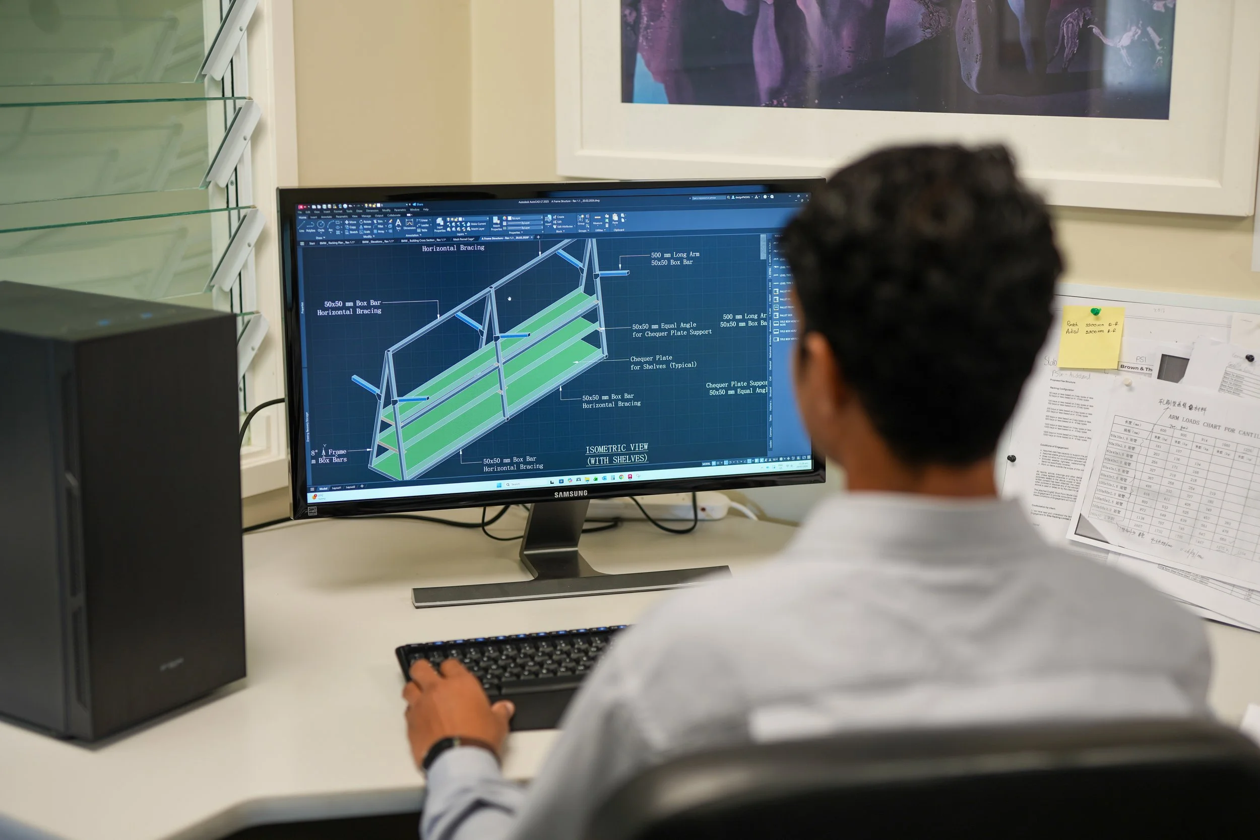 A person working on a computer with a technical 3D CAD design of a shelving unit on the screen.