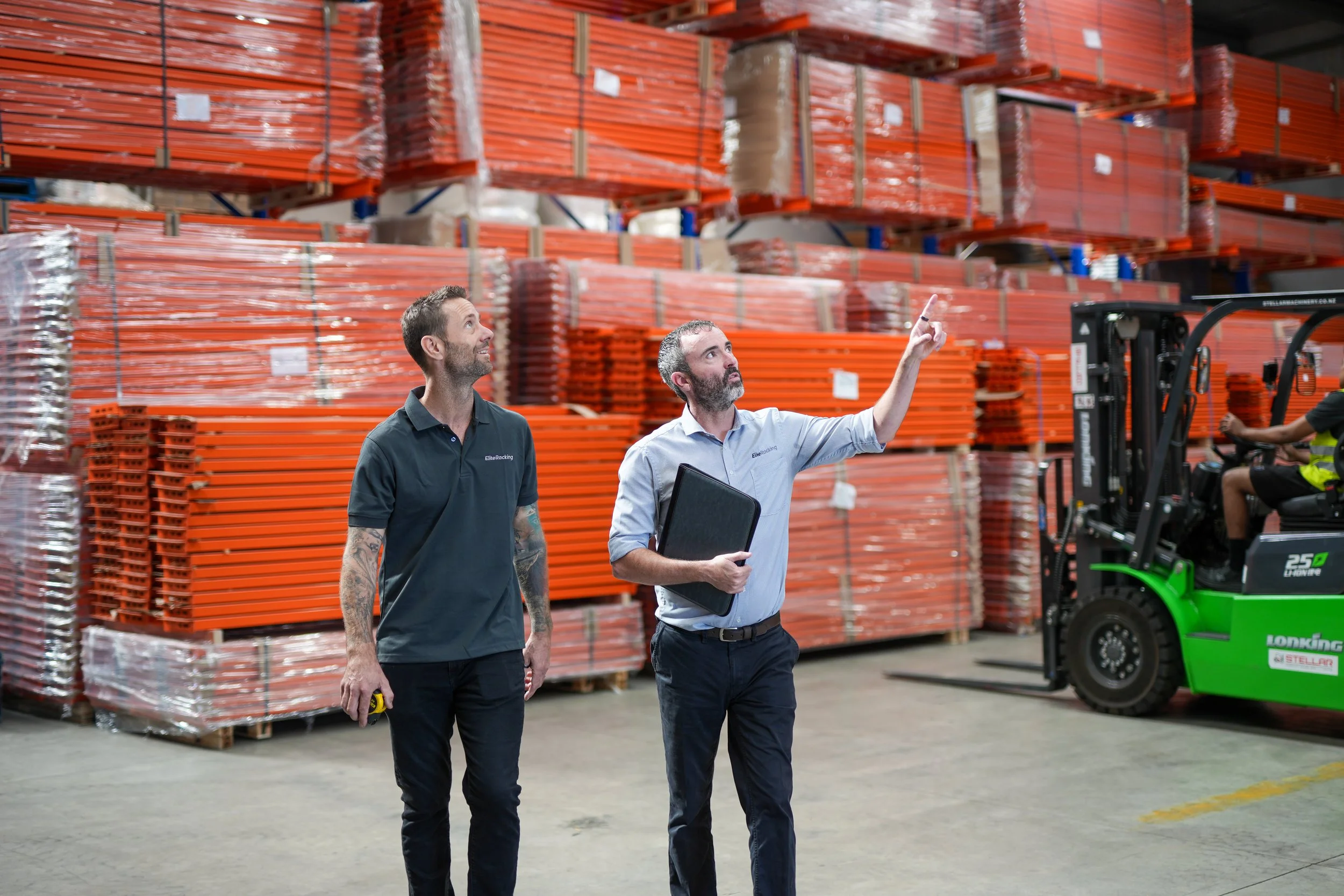 Two men are working in a warehouse surrounded by pallets of orange metal frames and a forklift. One man is pointing up while the other holds a binder.