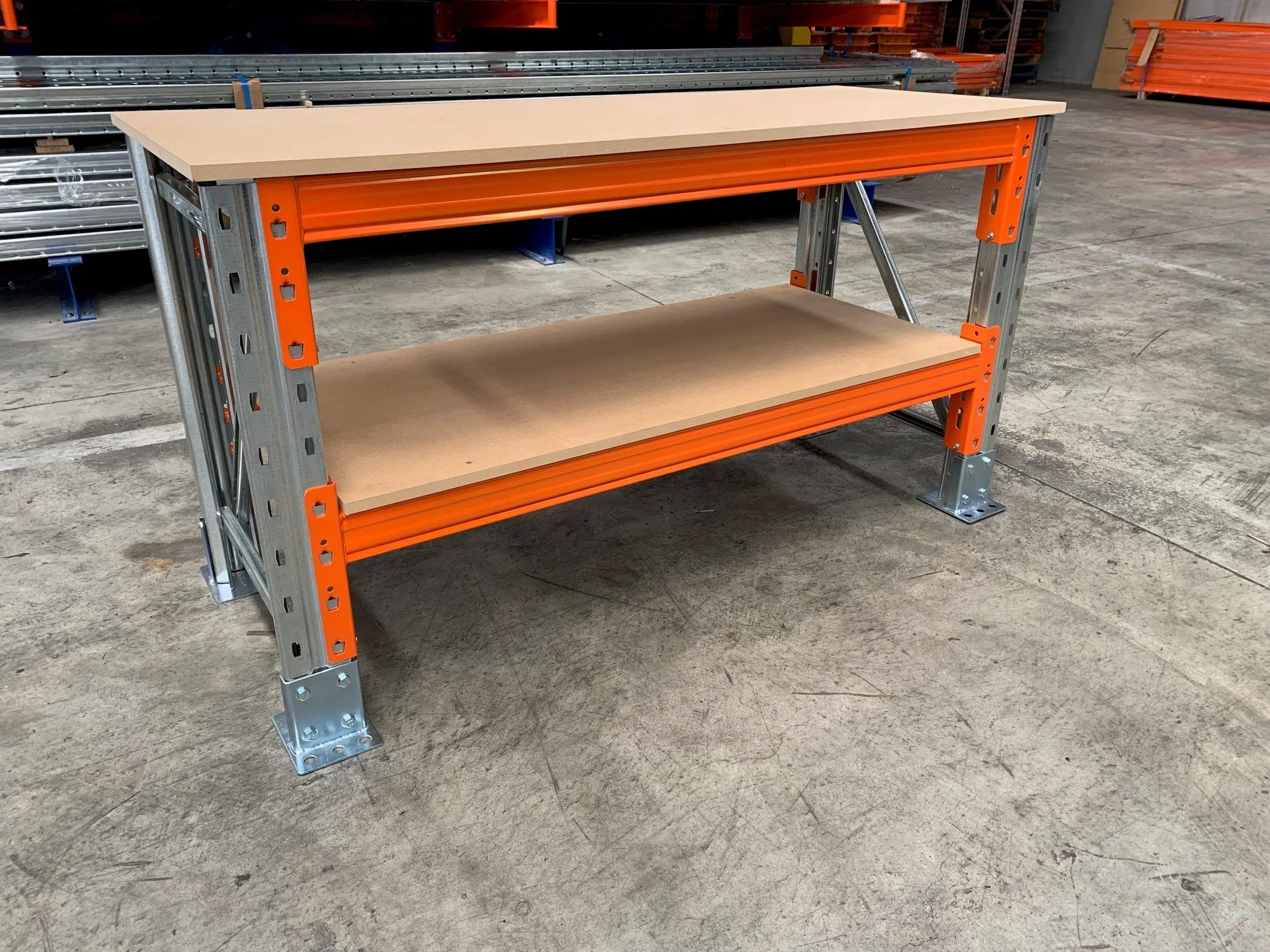 Industrial workbench with a beige top and a lower shelf, located in a warehouse with orange and metal shelving in the background.