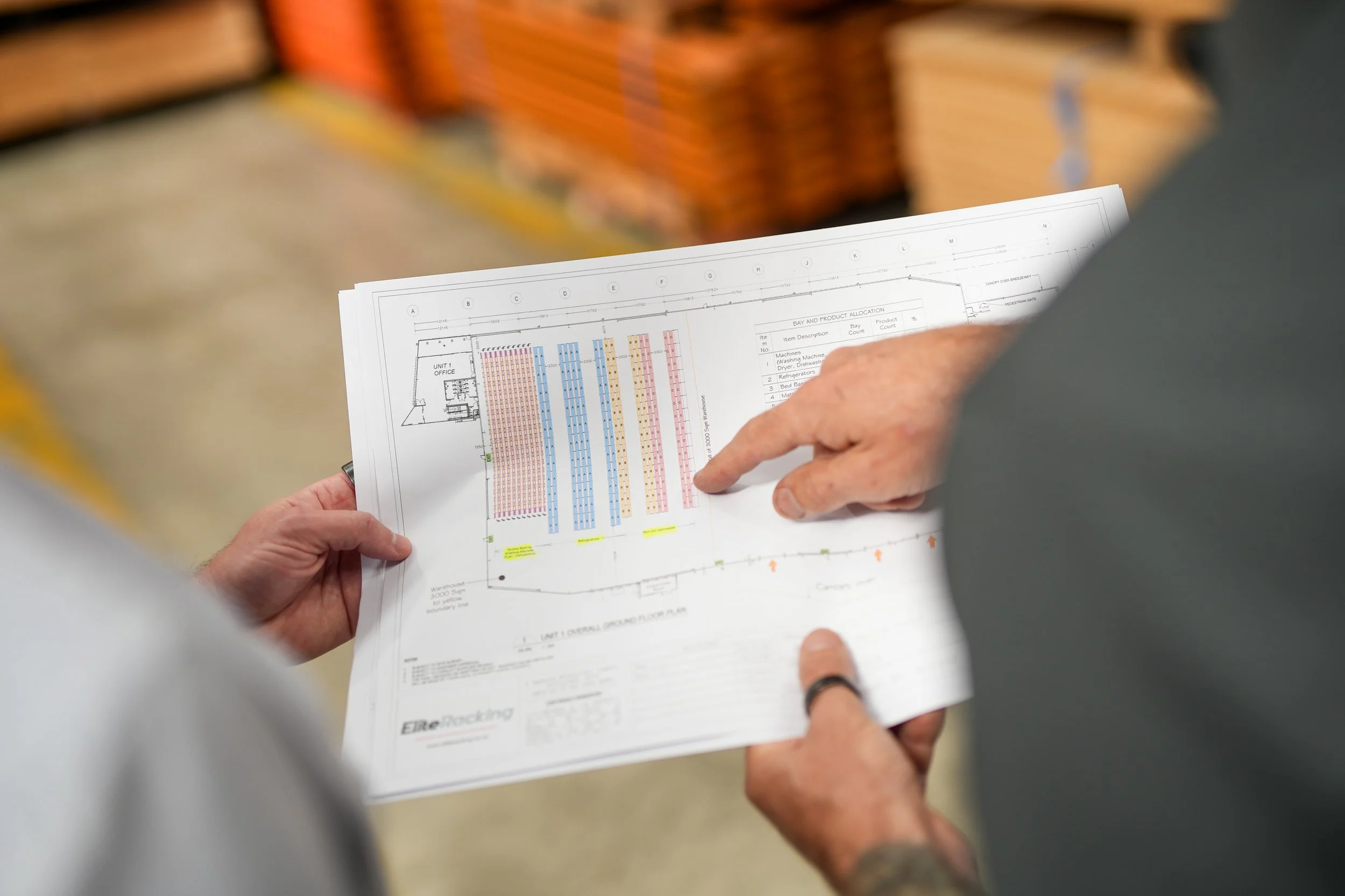 Two people examining a printed layout or floor plan, with colorful markings and highlighted sections, inside a warehouse or storage area.