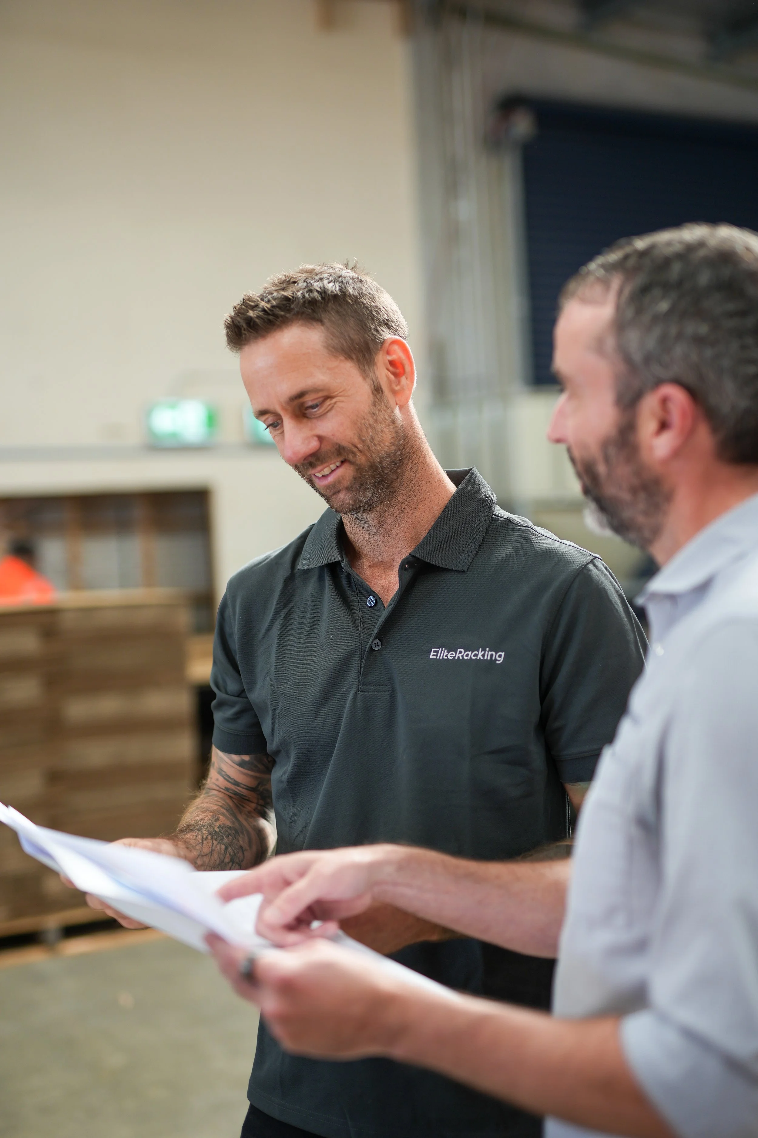 Two men having a conversation in an industrial setting, both holding papers and smiling.