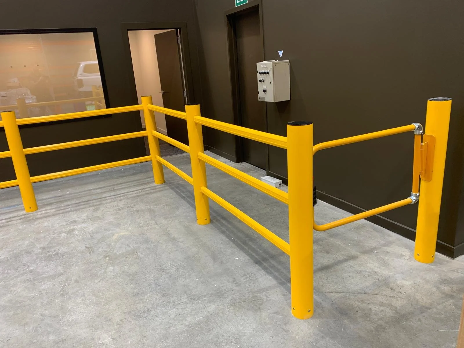 Bright yellow safety guardrail in an indoor industrial or commercial space with a dark wall, a door, and a window showing a vehicle outside.