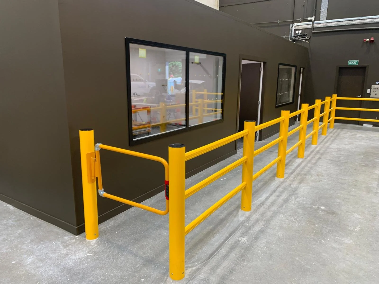 Yellow safety railings surrounding a dark-colored building with two windows and an exit door inside a parking area.