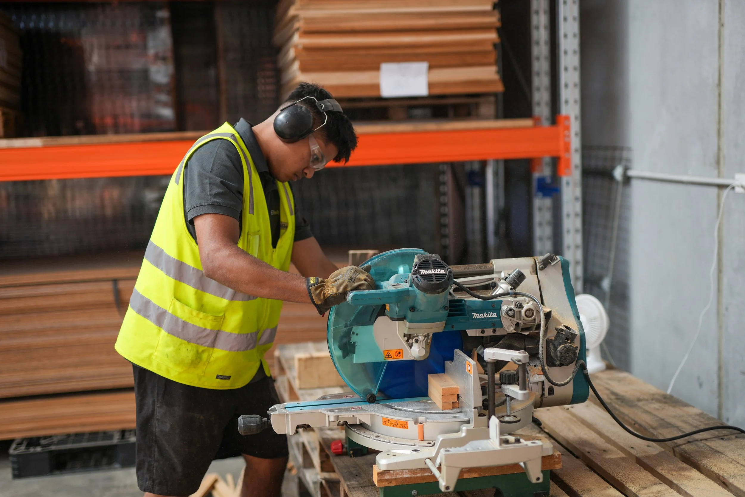 A man wearing safety glasses, earmuffs, gloves, and a yellow safety vest using a blue and white miter saw on a wooden workbench in a woodworking shop.