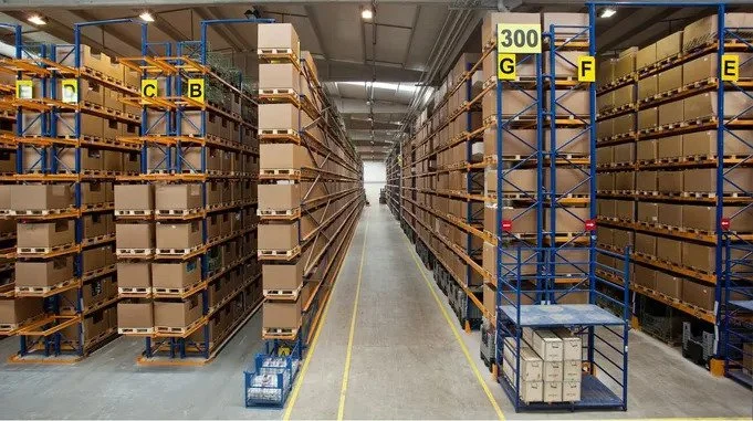 Warehouse aisle with tall shelving units filled with boxes, labeled with letters and numbers, and some pallets on the floor.